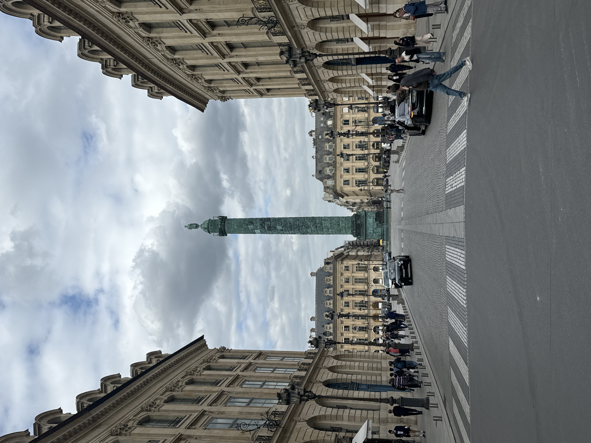The image depicts the Place Vendôme in Paris, France. In the center stands the Vendôme Column, a tall, bronze-covered monument. Flanking the square are symmetrical buildings with classical facades, featuring arched windows and detailed cornices. A street runs through the square, and people are walking on the sidewalks and crossing the street. Cars are also visible on the street. The sky above is overcast with cloudy skies.