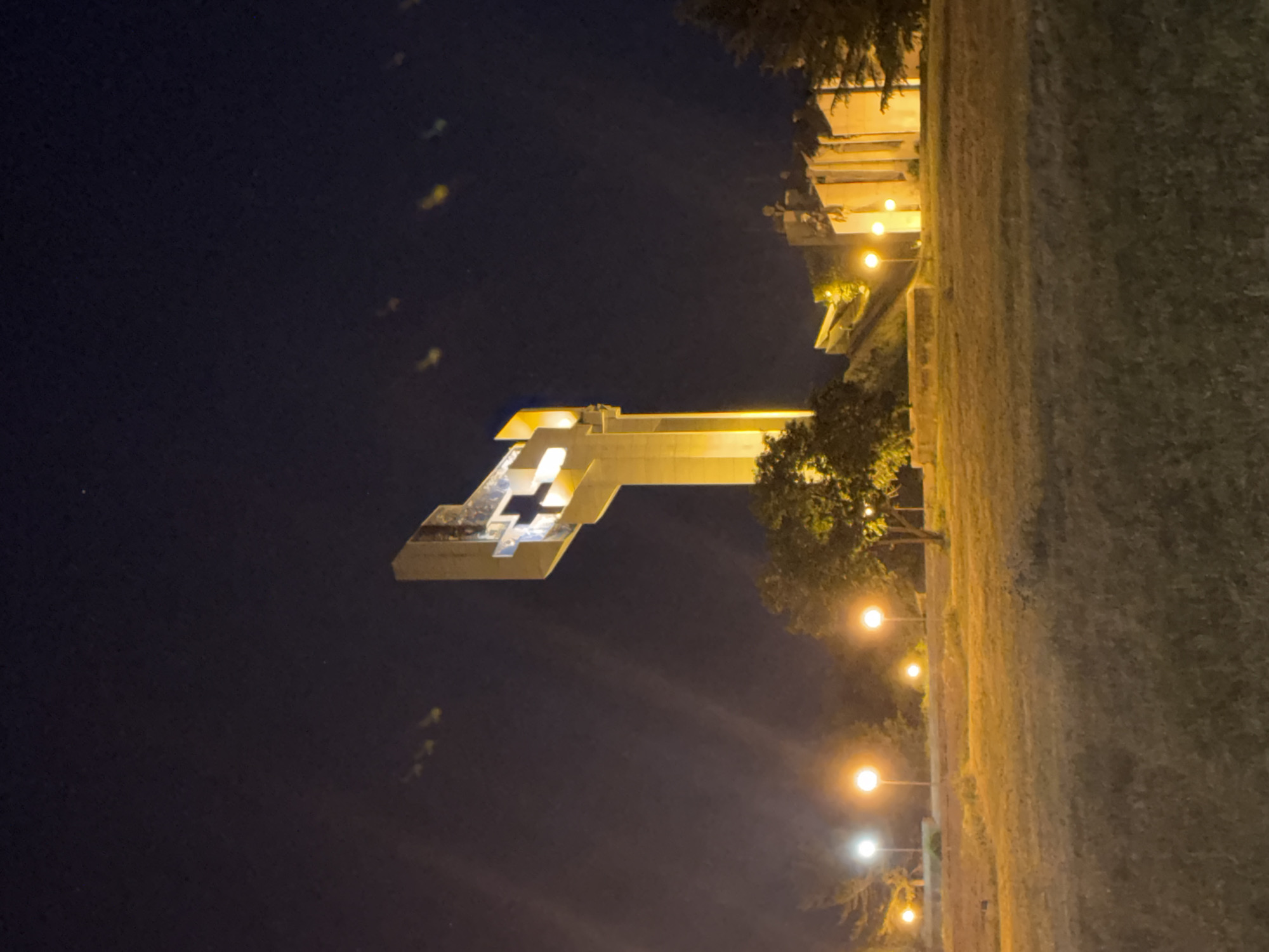 The image depicts the Defenders of Stara Zagora Memorial Complex at night, illuminated by artificial lighting. The monument, a large concrete structure with a cruciform opening, stands against a dark sky. A line of lampposts runs along the side, adjacent to a grassy area. Trees are interspersed throughout the scene. The image is oriented sideways. The location is Stara Zagora, Bulgaria. No people are visible.