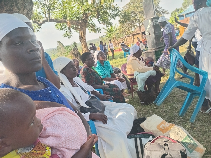 This image depicts an outdoor gathering, primarily of women and children, in a rural or semi-rural setting, consistent with Burundu, Kenya. The scene is illuminated by bright daylight, suggesting midday or early afternoon.
In the foreground, a woman wearing a blue top and a white head covering holds a baby wrapped in a pink blanket. Her expression appears serious or contemplative. She is seated on a blue plastic chair, as are several other women positioned behind and to her right. Many of these women also wear white head coverings and are dressed in various colored garments, including patterned dresses and solid blue or green tops. One woman further back in a yellow top is seen holding a child.
On the right side of the frame, a large black speaker with the text "ROYAL SOUND" and a partial phone number "075...19194" is prominent, suggesting music or announcements are part of the event. A woman in a purple patterned dress is standing near the speaker, while another person in a white top appears to be moving a blue plastic chair. Various bags, including a grey duffel bag and a white sack with "FRESH" printed in green, are visible on the ground in the lower right foreground.
The gathering takes place on a grassy area, shaded by large trees with green leaves. In the distant background, what appear to be simple village structures or huts can be seen. The overall impression is of a community or social event, possibly with a religious or cultural dimension, given the attire of many participants.