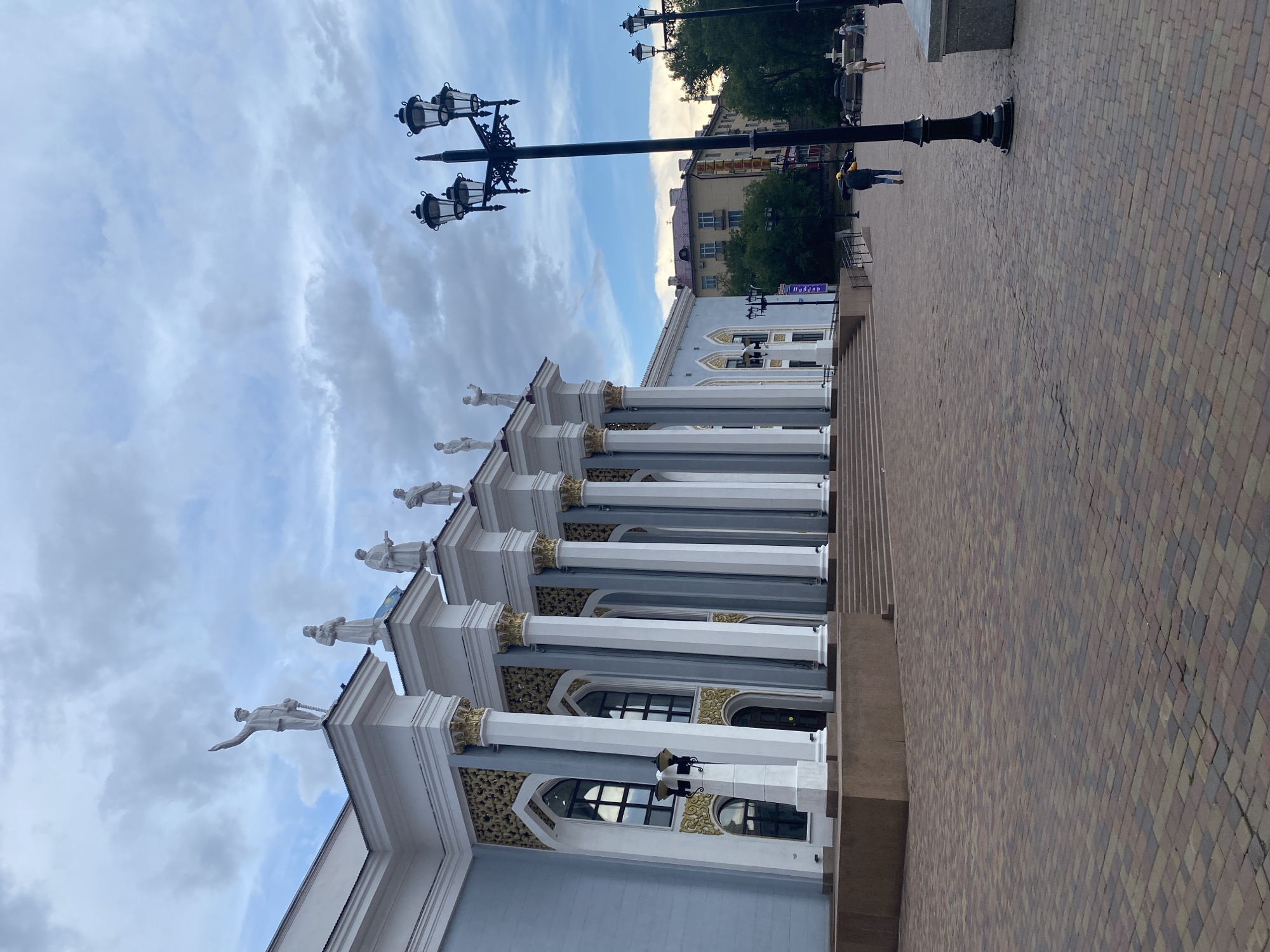 The image shows the Qarağandı city administration building in Kazakhstan. The building features a light blue facade with white columns and decorative gold accents. Several statues of figures in various poses are perched atop the building's roofline. A wide, paved plaza extends in front of the building, made of colorful bricks. Several ornate, black lampposts line the plaza. In the background, trees and other buildings are visible, as well as a few people walking. The sky is cloudy, with patches of blue showing through.