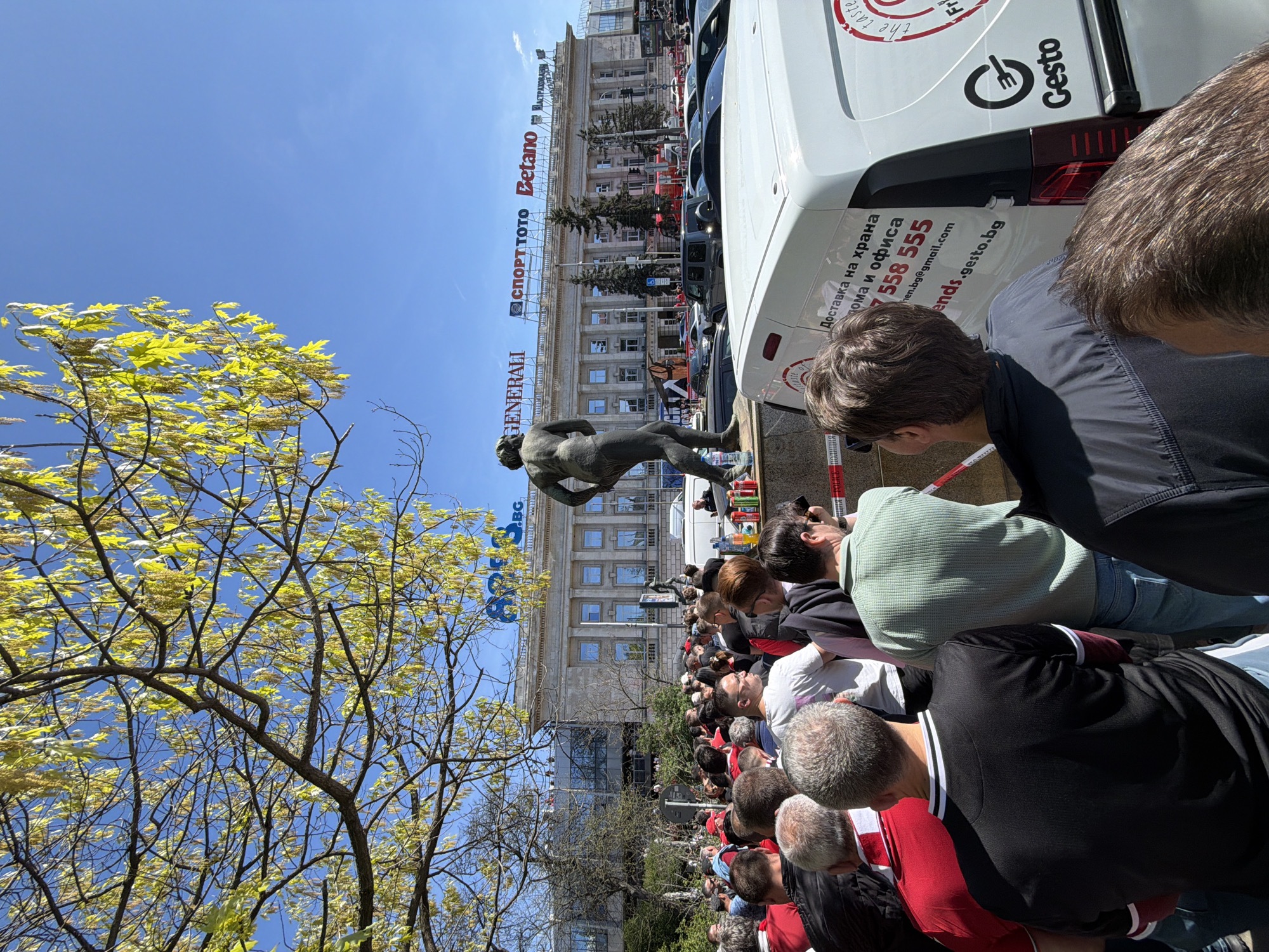 The image depicts a crowd of people gathered in front of a building. A sculpture of a male figure appears to be the focal point of the gathering. Banners with advertising logos are visible on the building's facade, identifying companies like "Generali," "Sport Toto," and "Betano." A van with the logo of "Gesto" is parked nearby. A tree with yellow-green leaves is visible on the left. The image provides visual information that implies that the scene is located in Sofia, Bulgaria, based on the Cyrillic script present on the banners and the van's livery. The crowd seems to be looking in the same direction.