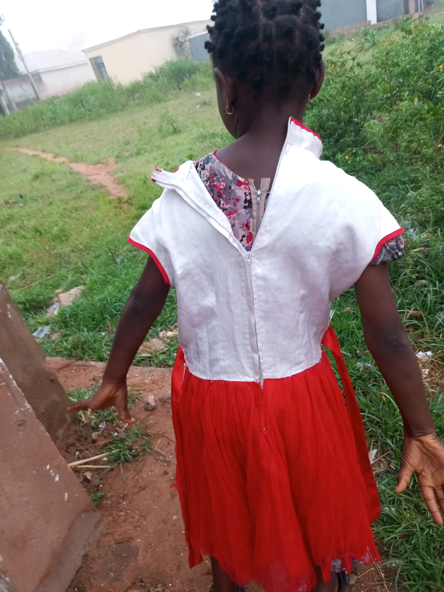 A rear view of a young Black girl wearing a red and white dress with a floral print is shown in this medium shot. The dress has a white bodice with short sleeves and a zipper down the back, while the skirt is a vibrant red. The girl has her back to the camera, and her arms are extended to her sides. She is standing outdoors in what appears to be a rural or semi-urban area, with dry dirt paths and overgrown green vegetation visible in the background. There are also some buildings in the distance. The lighting suggests it may be daytime, possibly overcast or late afternoon. The girl is wearing a gold hoop earring in her visible ear.