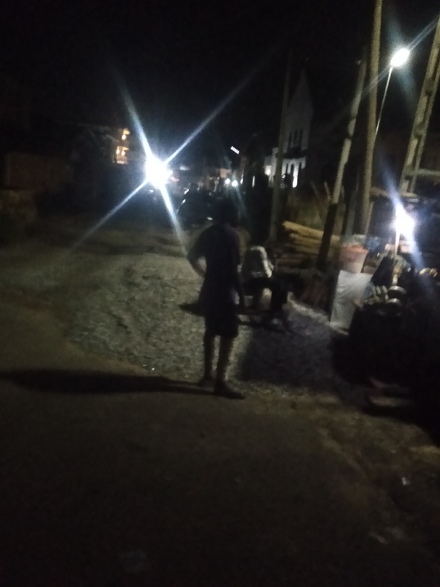 The image depicts a nighttime scene on an unpaved road or street in Shangisha, Nigeria. The environment is dark, illuminated primarily by artificial light sources that produce significant lens flares across the frame.
A prominent subject in the foreground, seen from behind, is a person wearing a dark t-shirt and shorts, standing on the rough ground. Their shadow extends forward onto the road. Further back on the right side of the frame, several other blurry figures are present, with one appearing to wear a light-colored top and possibly a cap. Adjacent to these figures are stacks of what seem to be wooden planks or logs, and a dark, indistinct object that might be a parked vehicle or equipment.
The background features several structures. On the left, a building with illuminated windows is visible in the distance. Towards the center-right, a distinct white building with a pointed or spired roof, possibly a church or similar institutional building, stands out. Multiple utility poles are visible on the right side of the road, with bright streetlights contributing to the overall illumination and lens flares. The road surface itself is dark, uneven, and appears to be composed of gravel or dirt.
The time of day is clearly night. No specific weather conditions are discernible, but the air appears clear enough for light to travel with bright intensity. No visible text is present in the image.