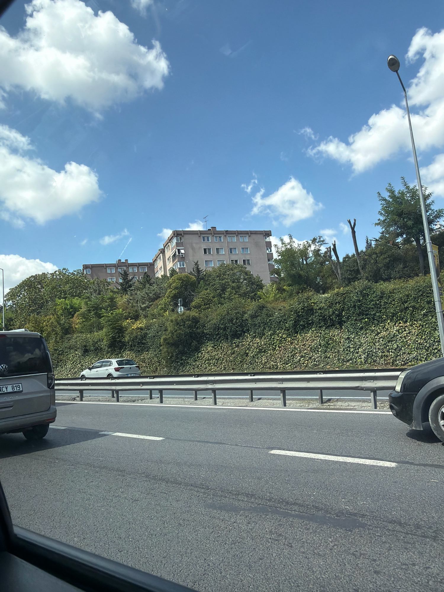 The image depicts a daytime scene from a vehicle's perspective, looking up a slope beside a highway. The sky is a clear blue with scattered white clouds. A tall lamppost is visible on the right side of the frame. On the slope, dense green foliage covers the ground, with trees and bushes growing higher up. Two apartment buildings, with multiple windows and balconies, are situated on the top of the slope, surrounded by trees. A white car is driving on the highway in the middle distance, and a gray van is in the foreground on the left. A black car is partially visible on the right foreground. The highway has clear lane markings. The scene is likely located in Bahçelievler, Turkey, given the context.
