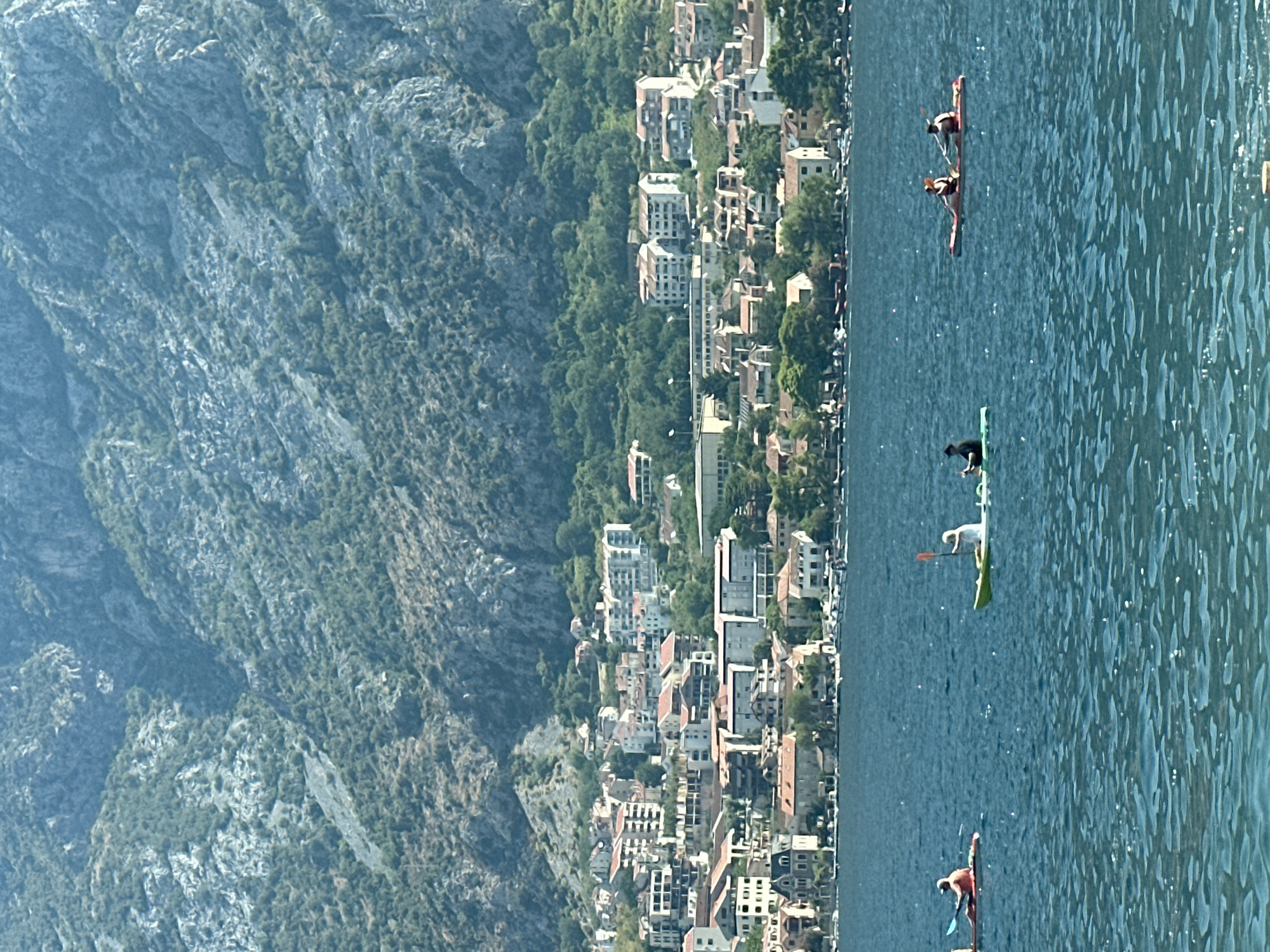 The image shows a scenic view of the Bay of Kotor in Montenegro, specifically depicting the town of Prčanj nestled at the foot of a steep, tree-covered mountain. The foreground is dominated by the calm, blue water of the bay, with sunlight reflecting off its surface. Several kayakers are visible on the water. On the left, a single kayaker in an orange kayak paddles away from the viewer. To their right, a lime green kayak carries two people, one of whom is holding a paddle. Further to the right, a red kayak is occupied by two individuals also holding paddles.
The mid-ground features the town of Prčanj, characterized by a dense collection of buildings with terracotta roofs, interspersed with lush green trees. The architecture appears to be a mix of older and more modern structures, including several multi-story apartment buildings. The town extends along the shoreline, with smaller boats docked or moored near the shore.
The background is dominated by the imposing mountain that rises sharply behind the town. The mountain's slopes are rugged and rocky, with patches of green vegetation, indicating a mix of bare rock and hardy plant life. The overall impression is of a tranquil, sun-drenched coastal scene.