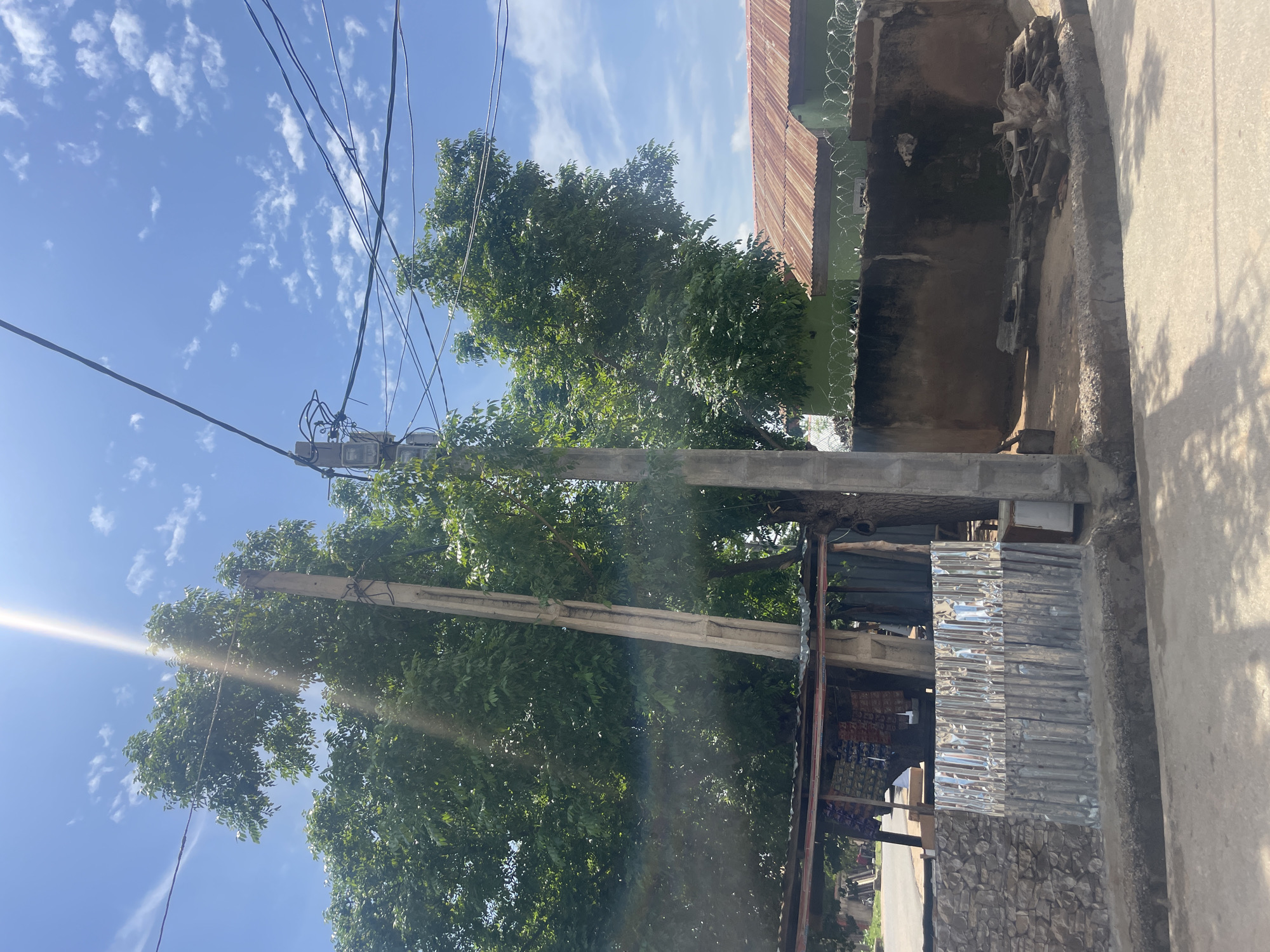 The image shows an outdoor scene, likely in Jimeta, Nigeria, on a sunny day with scattered clouds. The perspective is slightly tilted, looking upwards towards a utility pole adorned with electrical wires and transformers. A large tree with lush green foliage dominates the central part of the frame, its branches reaching towards the sky. In the background, a building with a rusty, corrugated metal roof is visible, topped with barbed wire. The building's wall appears to be made of concrete and has some staining. To the right, a concrete pathway or road runs alongside the building, casting shadows from unseen objects. Further to the right, part of another building's wall, also made of concrete, is visible. The lower part of the image shows some corrugated metal sheeting attached to a stone wall, possibly as a repair or modification to a structure. The overall impression is one of an urban or semi-urban environment, with a mix of natural elements and man-made structures. There are no people or explicit activities depicted. The bright sunlight suggests it is daytime, likely late morning or early afternoon.