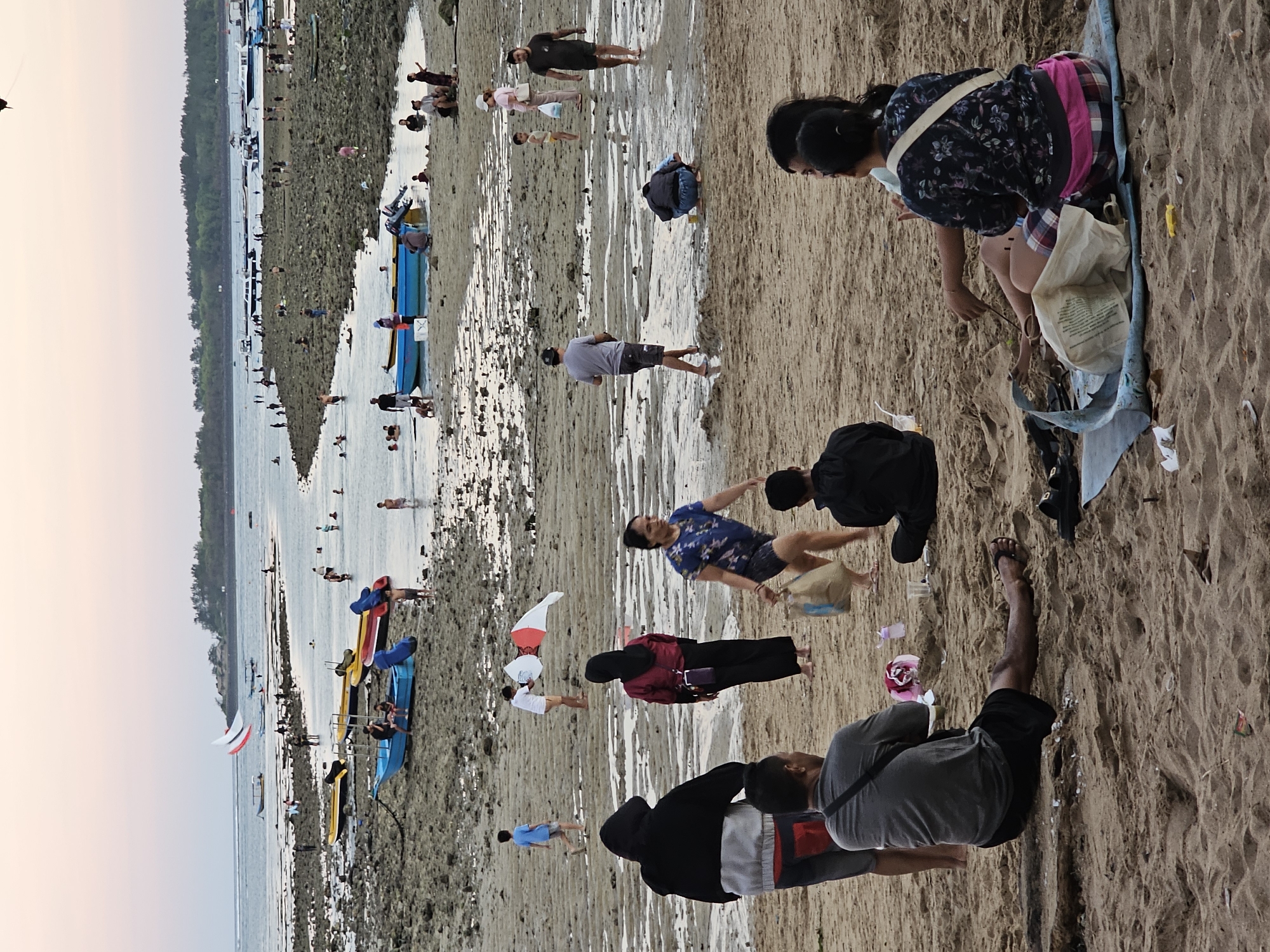 The image depicts a crowded beach scene in Denpasar, Indonesia, likely during low tide. The sand is wet and textured, with shallow pools of water reflecting the sky. Numerous people are scattered across the beach and in the shallow water. Some are walking, others are sitting or kneeling on the sand. Several colorful kayaks and small boats are pulled up onto the shore or floating in the water, suggesting recreational activities.
In the foreground, a group of people are gathered on the sand. One person, wearing a dark shirt and shorts, is sitting with their legs extended. Another person, wearing a grey shirt and black shorts, is also seated. A person with a black hoodie is partially visible. Further back, a person in a patterned top and pink skirt is sitting on a mat, looking down.
In the mid-ground and background, more individuals are visible wading in the water or walking along the shoreline. The sea extends to the horizon, with a line of trees forming a backdrop along the coast. Two kites, one white and red, and another white, are visible in the distance, adding a touch of color to the sky. The sky is a soft, pale color, indicative of either early morning or late evening light, possibly during sunset. The overall atmosphere appears relaxed and leisurely, with people enjoying the beach environment. There are no discernible signs or text in the image.