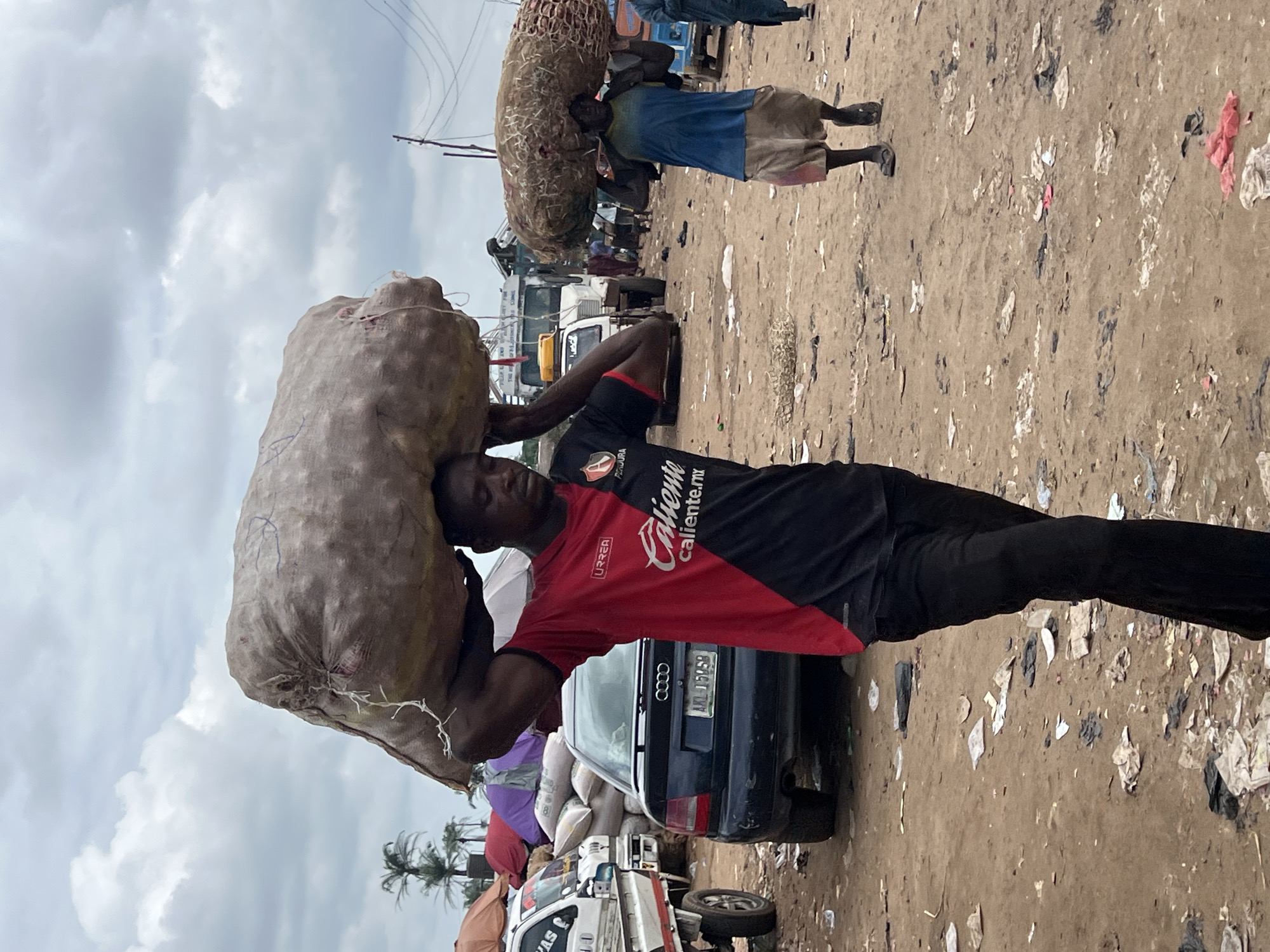 In Oko Amakom, Nigeria, under a cloudy sky, a man in a red and black shirt carries a large sack of produce on his head. He walks on a dusty, debris-strewn ground. Behind him, another man carries a similar sack on his head. The background shows a busy market scene with vehicles, including trucks and cars, and various stalls with umbrellas and goods. The environment is characterized by a dirt ground littered with trash and a general atmosphere of a bustling outdoor marketplace.