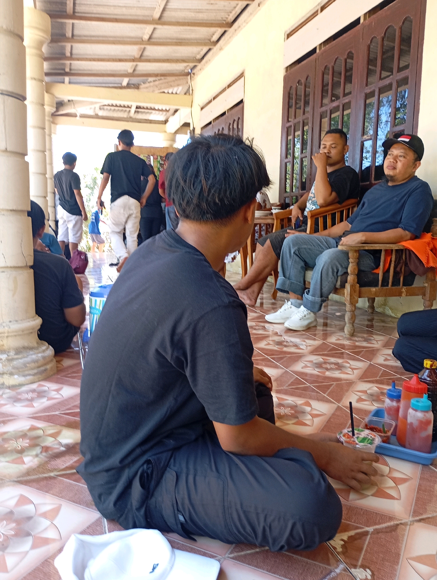 The image depicts a group of men gathered outdoors in a covered area, possibly a porch or veranda, in Ketepung, Indonesia. The scene is set on a tiled floor with a patterned design. In the foreground, a man in a black t-shirt and pants is seated on the floor, facing away from the camera. To his right, a tray with several condiment bottles and a cup with a straw is placed on the floor.
Further back, two men are seated on a wooden bench. The man on the left, wearing a black t-shirt, has his eyes closed and appears to be resting his chin on his hand. The man on the right, wearing a dark blue t-shirt and a baseball cap, is looking to his left and has his hands resting on his lap.
In the background, several other men are standing or walking. One man in a black t-shirt and white pants is walking away from the camera. Another man in a black t-shirt and white pants is standing further back. A glimpse of a red bag and a blue water cooler can be seen on the floor near the standing individuals. The environment suggests a casual, communal gathering.