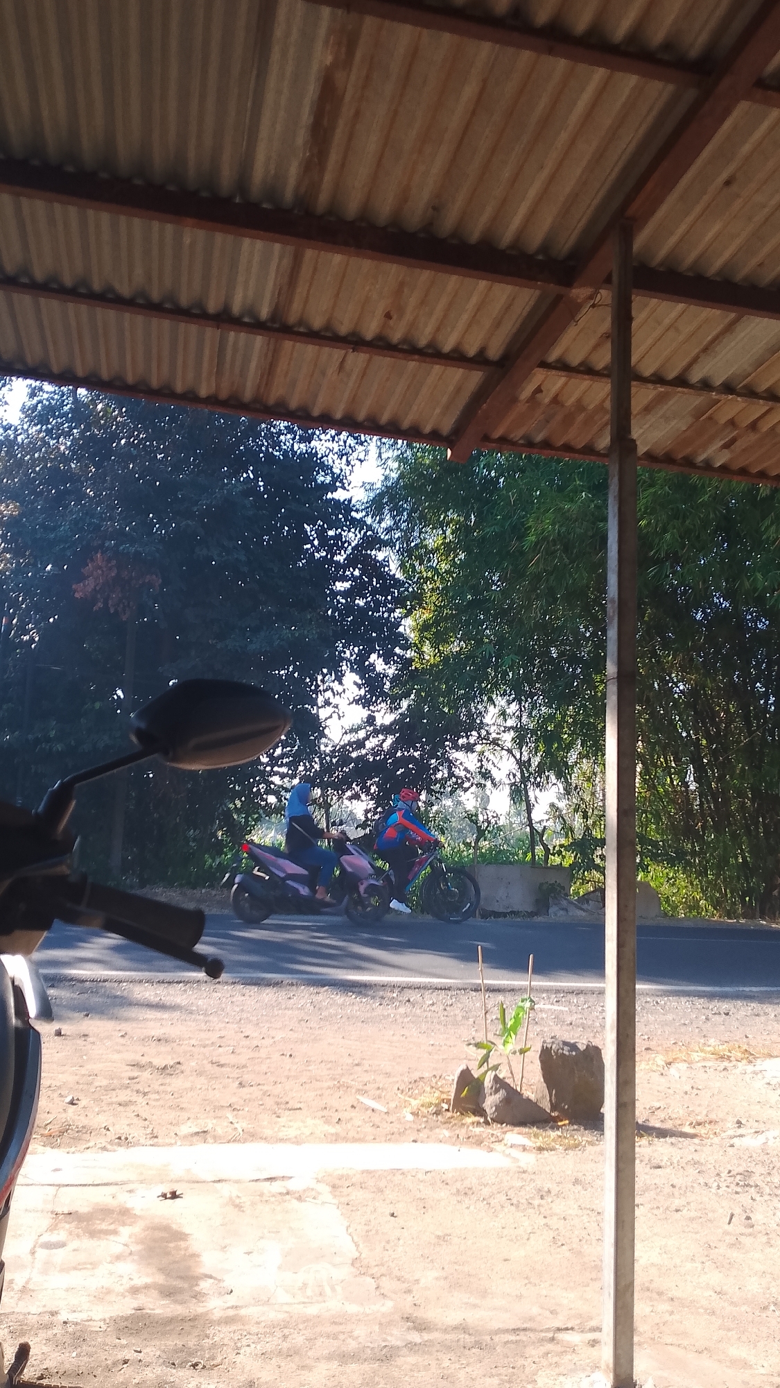 The image shows a view from under a corrugated metal roof, looking out towards a street. In the foreground, part of a motorcycle handlebar and mirror are visible on the left. Two motorcycles are on the street, with riders. The motorcycle on the left is pink and a person wearing a blue headscarf and dark clothing is riding it. The motorcycle on the right is darker and a person wearing a red and blue helmet and a blue and red jacket is riding it. There are trees and bamboo to the right of the street. The ground in the foreground appears to be a mix of dirt and concrete. The scene is set in a sunny outdoor environment.