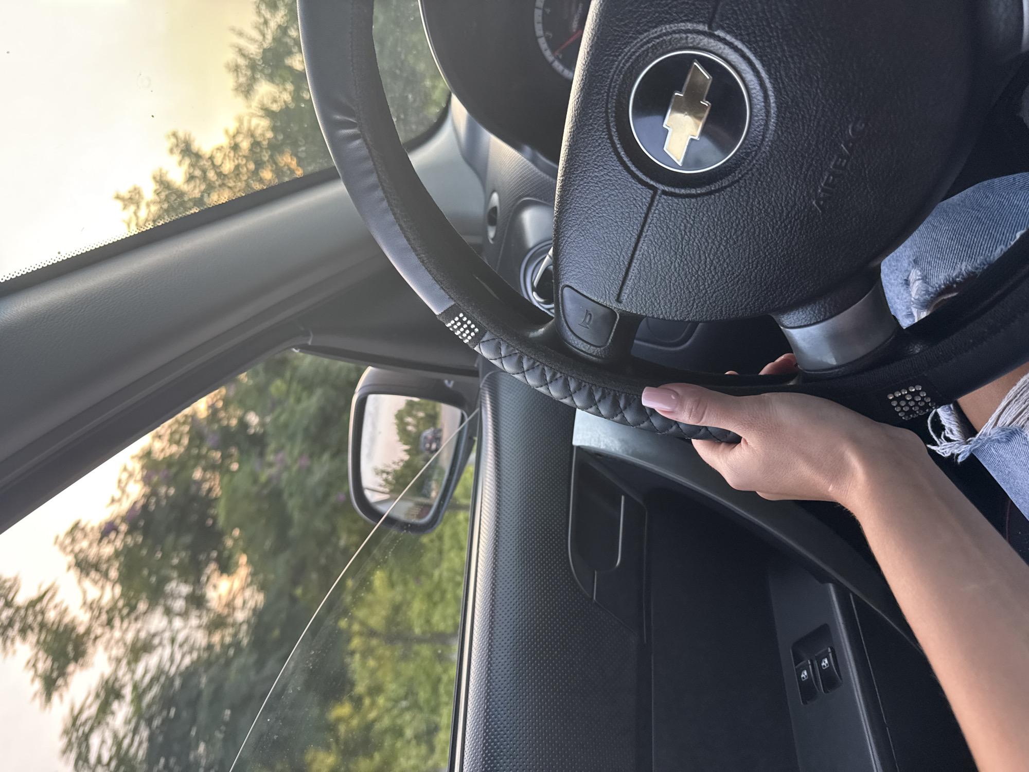 The image shows a close-up view of a person's hand and arm on the steering wheel of a car. The hand, with manicured fingernails, is gripping the black leather-wrapped steering wheel, which has decorative rhinestones. The car's interior, including the dashboard and door panel, is visible, along with the Chevrolet logo on the steering wheel. Outside the car, through the window, a blurry background of green trees and a bright, hazy sky suggests a daytime setting. The scene is likely captured while driving, as indicated by the hand on the wheel and the view of the road and surrounding environment in the side mirror. The location is identified as Cimișlia, Moldova.