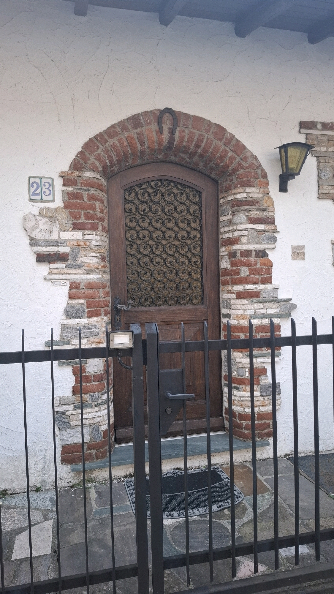 A low-angle, full shot shows a rustic arched doorway in an off-white stucco wall. The arch is constructed from red bricks, and a dark metal horseshoe is affixed to the top of the arch. The wooden door has a square inset panel with intricate metalwork. To the left of the door, a square ceramic tile with the number "23" is mounted on the wall. To the right of the door, a black lantern with a yellow glass pane is attached to the wall. The wall around the door is decorated with a mosaic of exposed bricks and flat stones, creating a textured, broken appearance. A black metal gate with vertical bars stands in front of the door. A doormat with a decorative border and the word "WELCOME" is placed on the stone-tiled ground in front of the door. The ground is paved with irregularly shaped stones in shades of gray, brown, and green. The scene appears to be an exterior view of a building entrance.