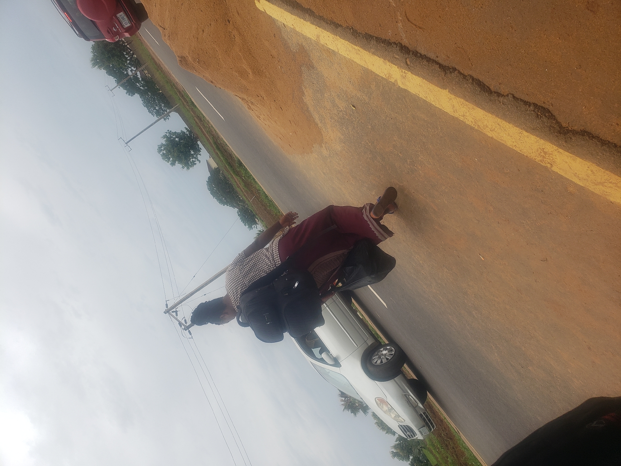 A woman, seen from behind, walks along a paved road in Gumel, Nigeria. She wears a patterned shirt and a maroon skirt, and carries a black backpack and a black plastic bag. A silver sedan is parked on the left side of the road, and a red SUV is visible further down the road on the right. The sky is overcast, and trees and power lines are visible in the background. The ground is dusty and brown, with a yellow line painted on the road.