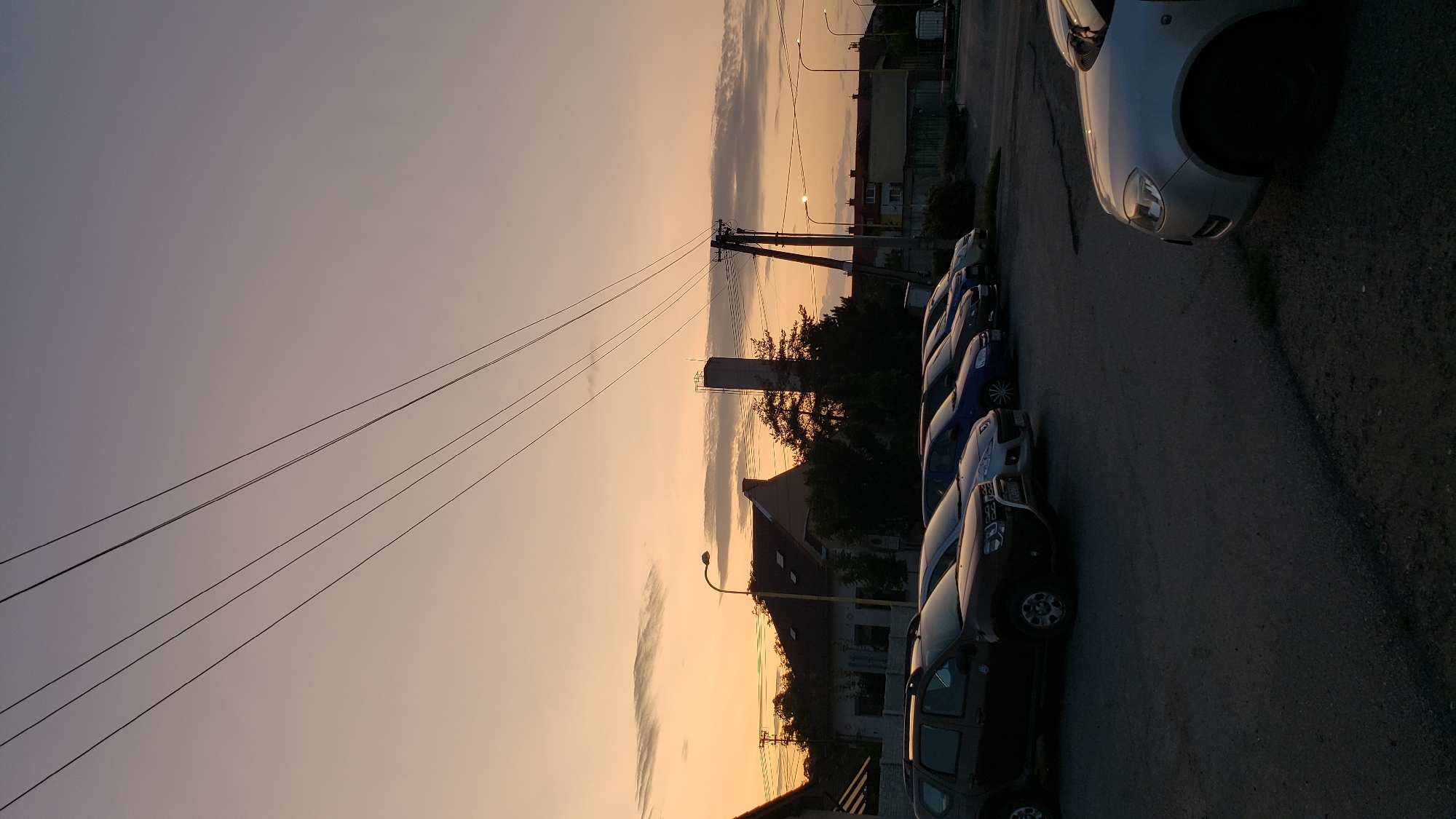 The image depicts a street scene at dusk in Prešov, Slovakia. A row of parked cars is visible on a paved surface, with buildings and trees in the background. Overhead, power lines stretch across the sky, which is tinged with the warm colors of sunset. A solitary lamppost stands to the left of the frame. The general atmosphere is quiet and still, suggesting a residential or commercial area.