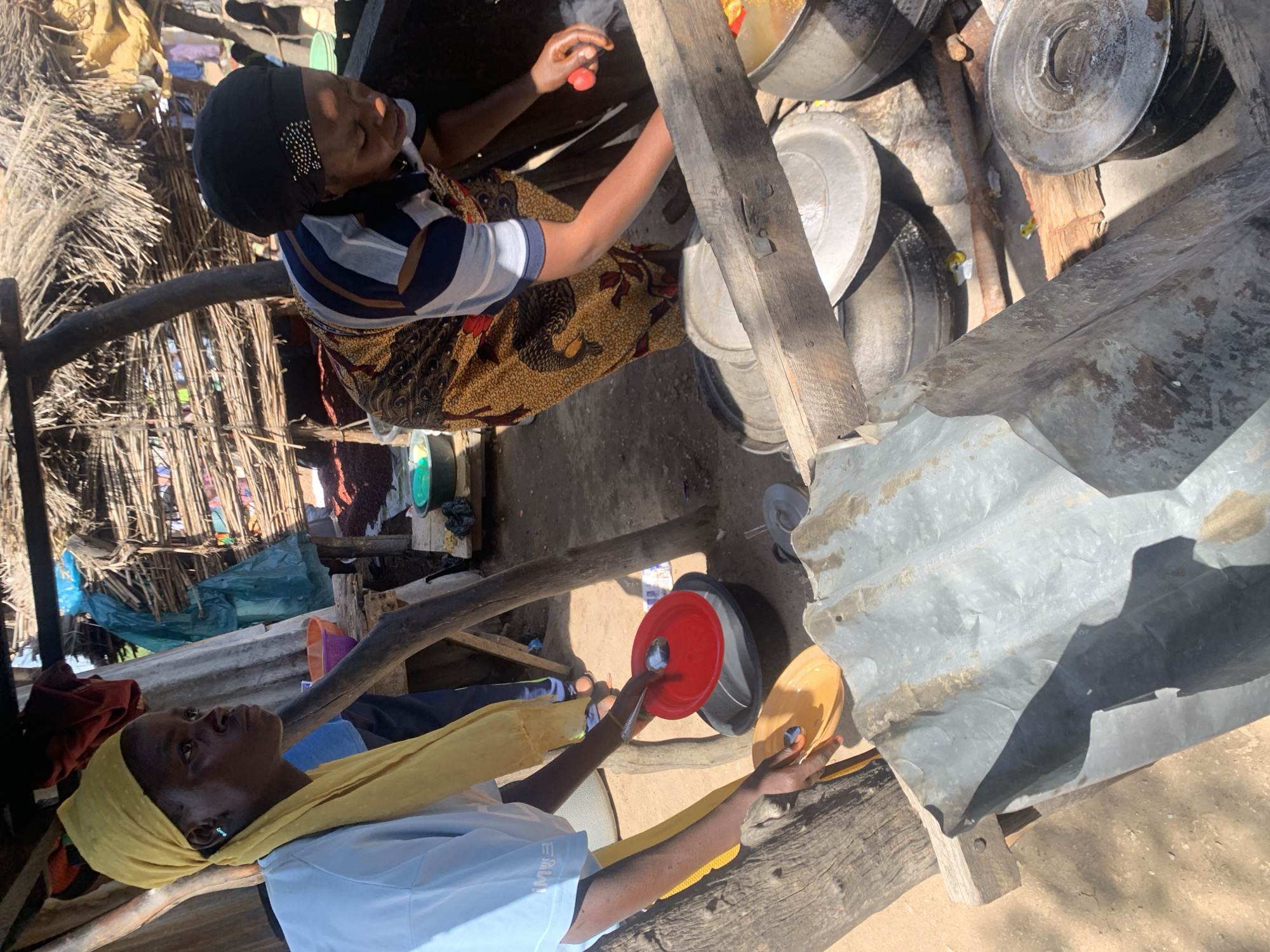 The image captures two individuals engaged in what appears to be food preparation in an outdoor setting. The scene is rustic, with a thatched roof structure and rough wooden supports suggesting a rural or informal dwelling.
In the foreground, a person wearing a yellow headscarf and a light blue shirt looks upwards, their head tilted back. Their right hand is visible, holding a spoon over a red plate. Next to the red plate is a yellow plate. In the background, another person, partially visible, wears a black head covering and a striped shirt. They are attending to a large pot, with their hands near what looks like a tomato. Around them are several large metal pots and containers, some with lids. The ground is dirt, and sunlight casts shadows, indicating it is daytime, likely outdoors. The overall atmosphere is one of daily life and communal activity. There is no visible text in the image.