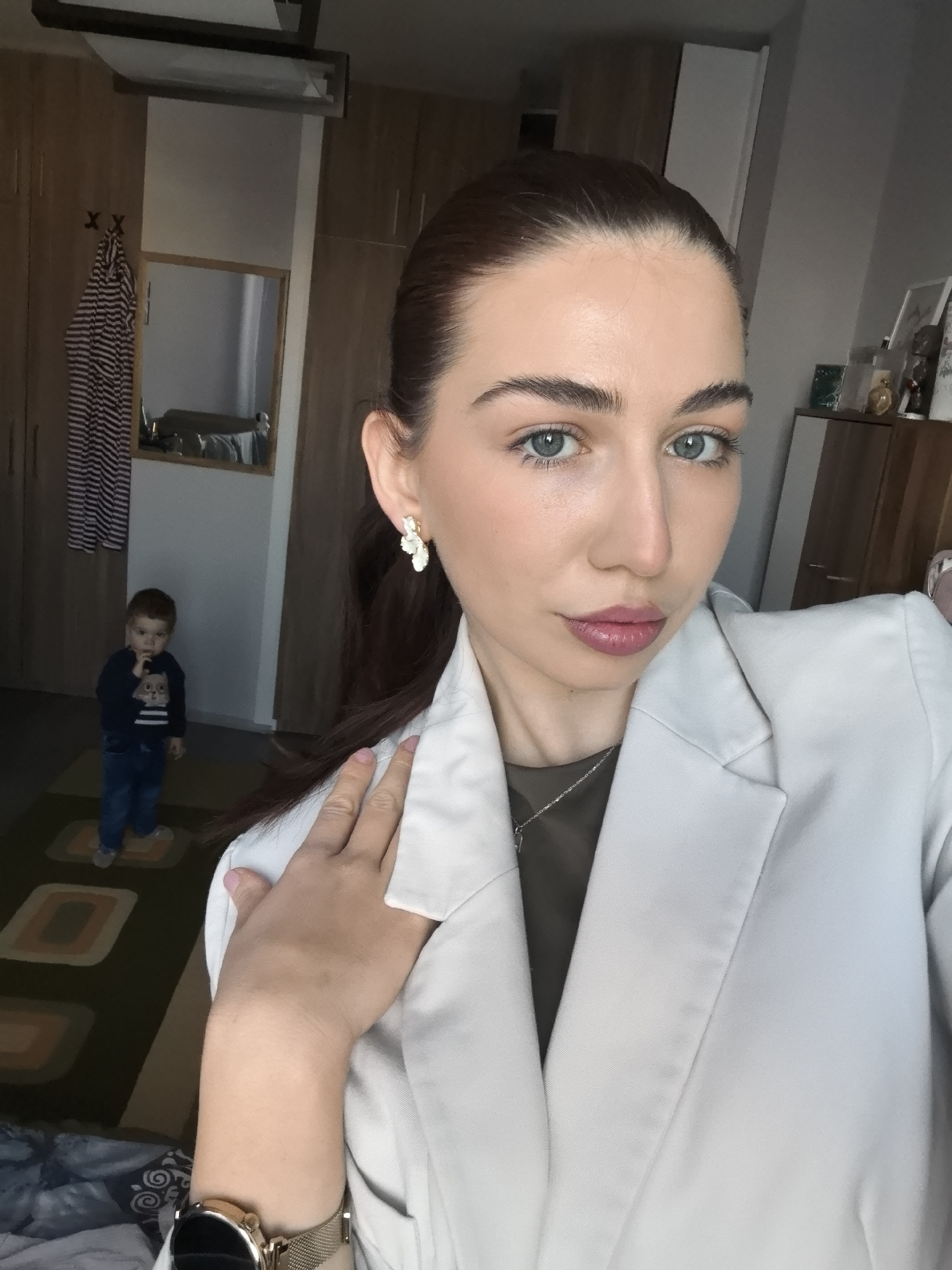 The photograph shows a woman in a light-colored blazer, wearing a necklace and floral earrings, in an indoor setting. She has fair skin, blue eyes, and dark hair pulled back. A child, wearing a navy sweater and jeans, is visible in the background near a doorway. A striped garment hangs on the back of the door. In the background there are wooden cabinets and a mirror. The location is likely in Stara Zagora, Bulgaria, based on the presence of a child who likely lives in the area, the style of the furniture, and the lack of any specific indicators that would place the image elsewhere.