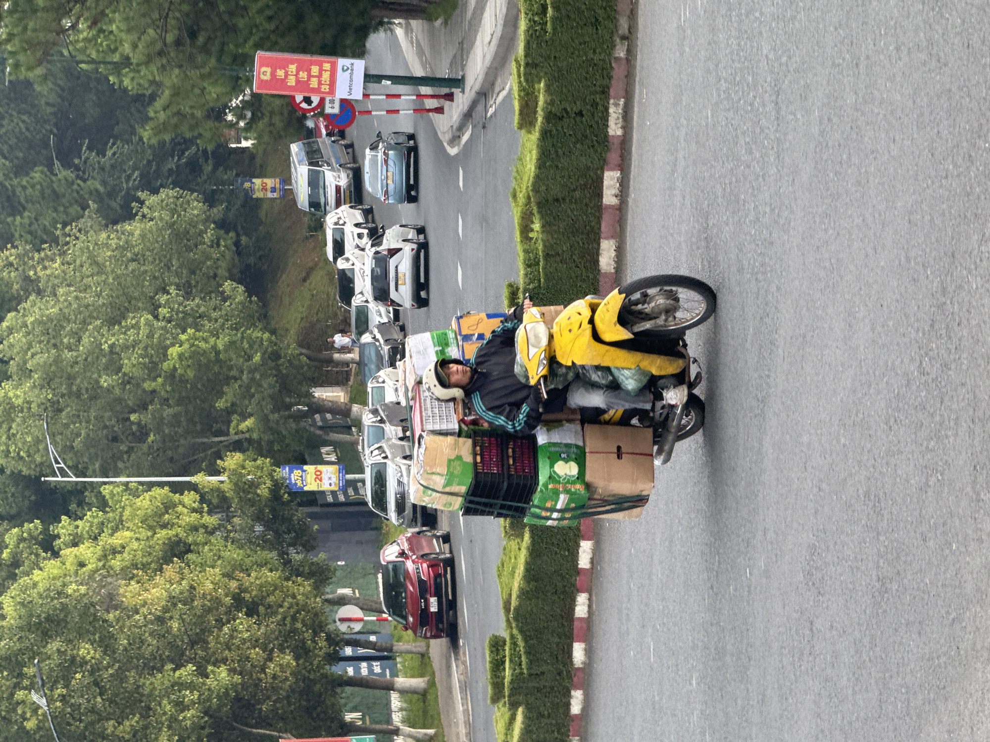 The image shows a person riding a yellow scooter loaded with many boxes and crates through a street in Da Lat, Vietnam. The person is wearing a black jacket and a white helmet. The scooter is heavily laden with various items, including stacked cardboard boxes and plastic crates, some of which are green and black. In the background, there are cars, vans, and a bus on the road, suggesting a busy urban environment. Lush green trees line the sides of the street, and there are hedges and streetlights visible. A red car is parked on the left side of the road, and a few traffic signs are present. The scene is captured in daylight, with a slightly overcast sky.