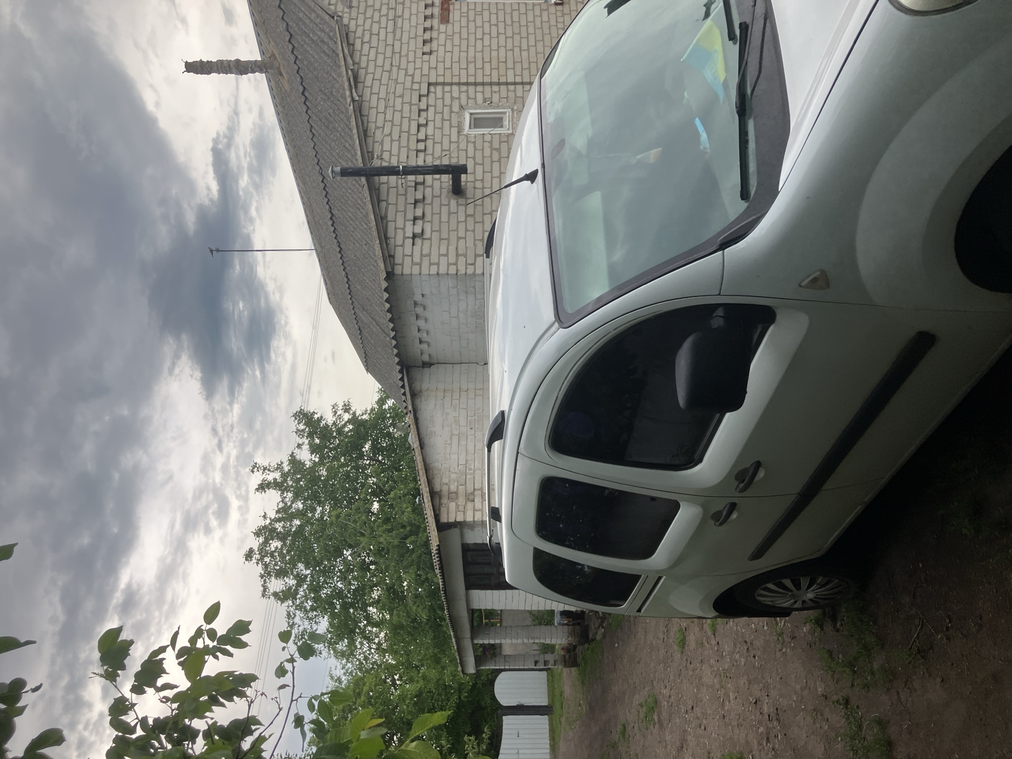 The image shows a white panel van parked on a dirt road in front of a brick building with a corrugated roof. Trees and foliage are visible in the background, along with a white fence. The sky is overcast with dark clouds. A chimney extends from the roof of the building. The van has roof racks and tinted windows. The scene appears to be in a rural or suburban area. The location is Vovkivka, Ukraine.