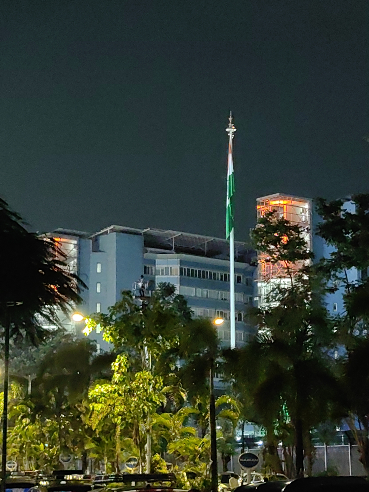 The image captures a dimly lit night scene in Mumbai, India, with an Indian flag prominently displayed on a tall flagpole in the foreground. The flag, with its distinct saffron, white, and green bands and the Ashoka Chakra in the center, stands against a dark sky. Behind the flag, a multi-story building with a light blue facade and numerous windows is visible. Some of the windows appear illuminated, suggesting occupancy.
To the right of the flag and the blue building, another structure rises higher. This structure is partially covered in scaffolding and glows with an intense orange light, possibly indicating construction or renovation work. Trees with lush green foliage dot the landscape in front of the buildings, and some streetlights cast a warm glow on the vegetation. In the lower part of the image, the silhouettes of several vehicles, possibly cars, are visible, suggesting a public or commercial area. A person can be seen on a platform or balcony of the blue building, near the flagpole. The overall atmosphere is one of urban nighttime activity, with a blend of national pride represented by the flag and the ongoing development or illumination of the city.