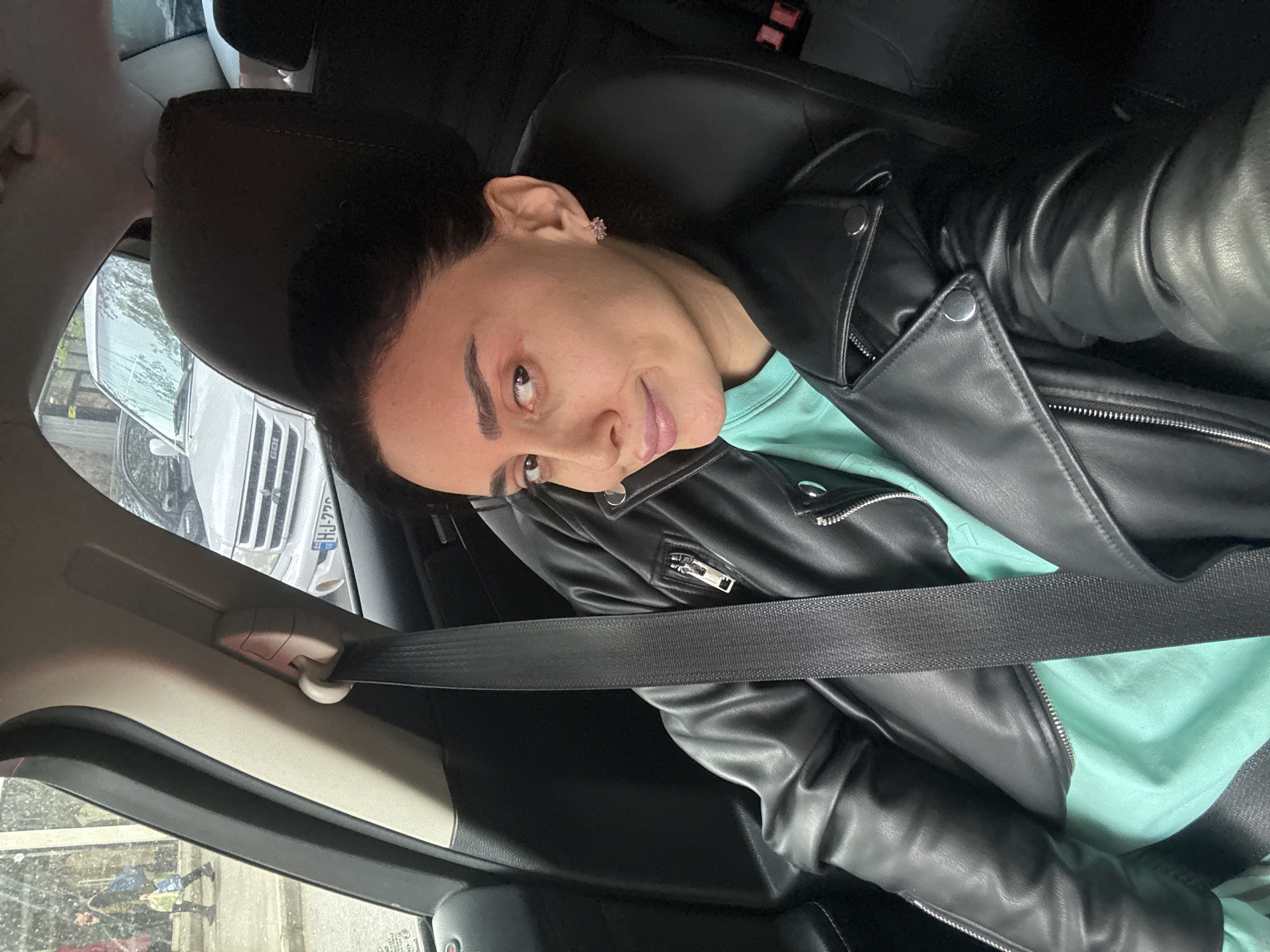 A woman is depicted inside a car, likely the front passenger seat, angled towards the camera in what appears to be a selfie or posed photo. She has dark hair pulled back, light brown eyes, and dark, well-defined eyebrows, with a faint smile or smirk on her face. She is wearing a black leather jacket over a mint green top and a prominent pink, flower-shaped earring in her right ear. A black seatbelt is visible across her chest and lap, indicating she is belted in.
The setting is the interior of a vehicle, featuring black leather upholstery on the seat and headrest. Through the car window, an outdoor scene is visible. Part of a white vehicle, possibly an SUV or truck, with a silver grille is seen. Reflections on the window suggest an urban or street environment, with a blurry reflection of a person in a light-colored jacket also visible.
Notable details include the crisp texture of the leather jacket and the vibrant color of her top. The white vehicle outside the car has the partial text "GDI" on its grille and "UJ-770" visible on its license plate, which includes Georgian characters "UJ." The lighting suggests it is daytime, possibly overcast, given the subdued light and reflections. The location context of Tbilisi, Georgia aligns with the Georgian license plate characters.