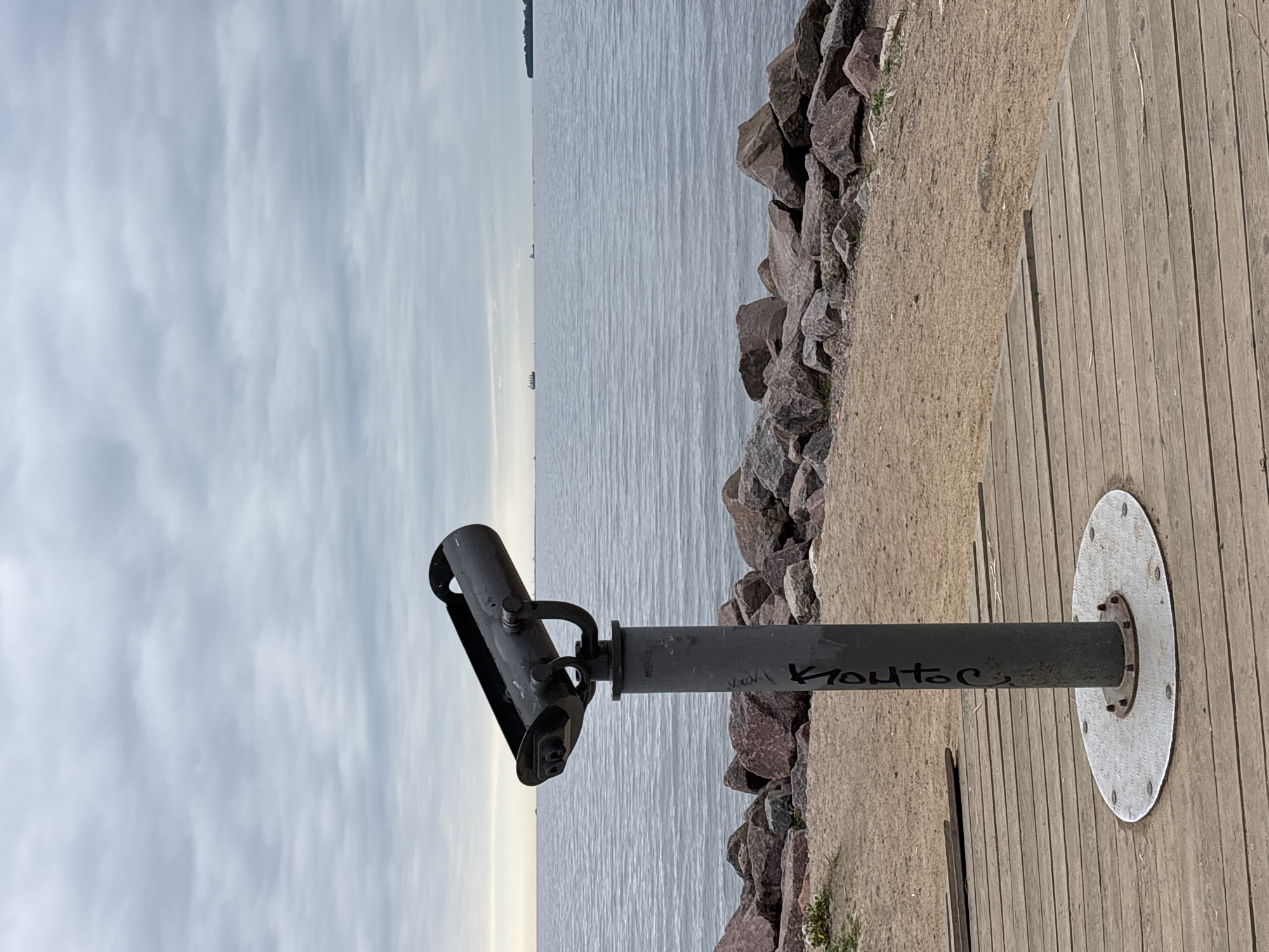 A medium shot presents a fixed binocular telescope standing on a wooden boardwalk, set against a backdrop of the sea and sky. The binocular telescope, centrally positioned, is made of metal and shows some graffiti on its support pole. The boardwalk stretches from the right edge of the image, made of light brown wooden planks. To the left of the boardwalk is a stretch of sand with scattered rocks. Beyond this lies a calm, gray sea. A few distant ships are visible on the horizon. The sky is overcast with a mix of white and gray clouds. This scene is located in Saint Petersburg, Russia.