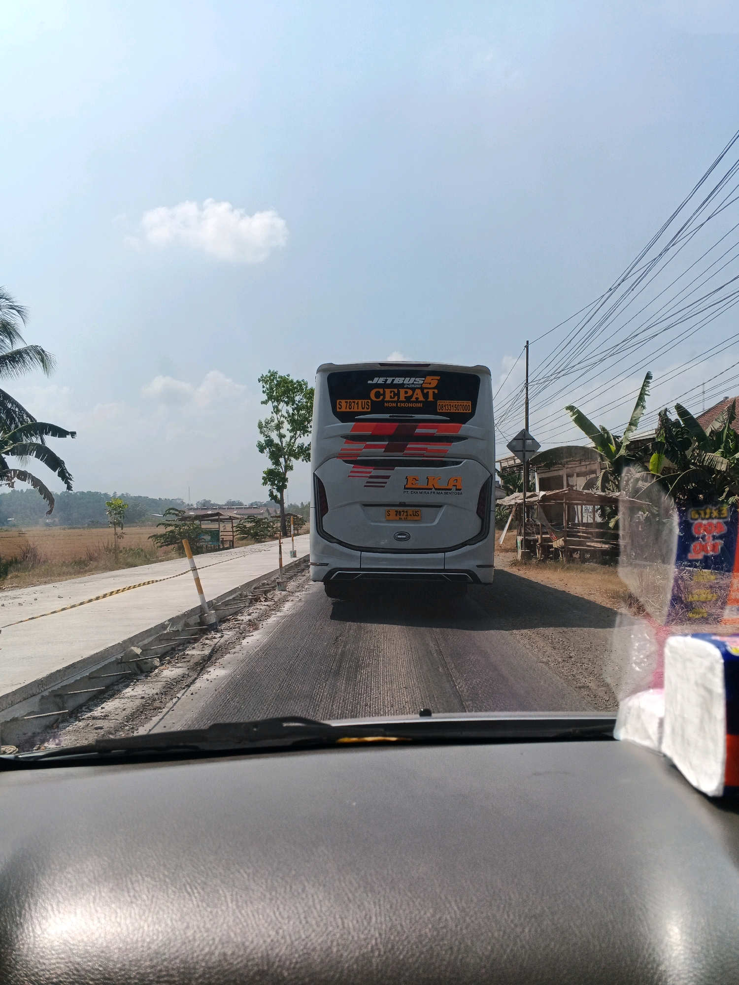 The image captures a view from inside a vehicle, looking forward at a road in Kedungpring, Indonesia, on a clear, sunny day. In the foreground, the dashboard and windshield of the vehicle are visible, with a packet of tissues on the passenger side. The road ahead is paved, with a construction site visible on the left, where new concrete barriers are being installed. A white bus with "JETBUS CEPAT" branding and a license plate starting with "S 7871 US" is traveling on the road. The background features rural scenery with trees, fields, and some scattered buildings. Power lines are strung across the sky on the right side of the image. A single, fluffy white cloud is present in the bright blue sky.