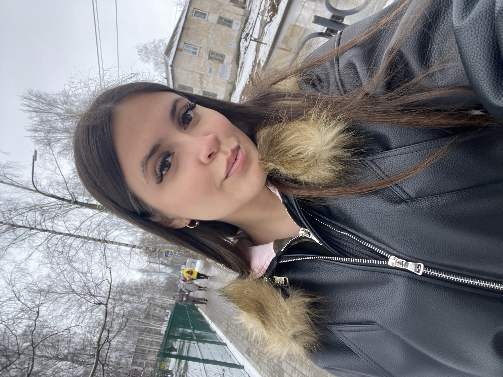 The image presents a selfie of a young woman. She has long, dark brown hair and fair skin. She is wearing a black leather jacket with a furry collar and a light pink shirt underneath.
The background shows an urban environment, likely in winter, due to the presence of snow. Bare trees and a building are visible in the background. There are a few people walking on the sidewalk. The presence of snow suggests a cold climate and helps place the location in a region with winter weather.
Given the specification that the location is in the city of Chaikovsky, Russia, the buildings in the background appear to be consistent with urban architecture in that region.