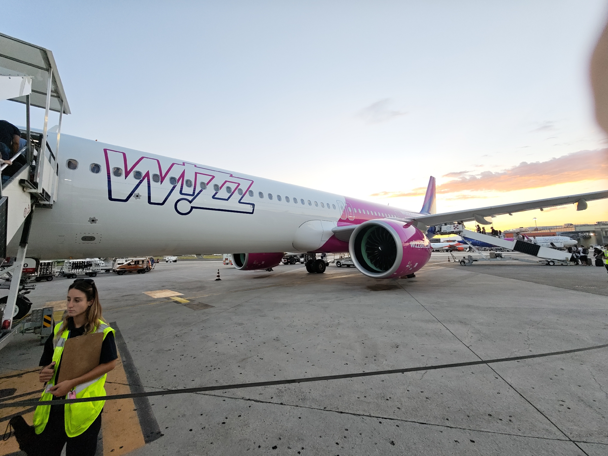 An airline employee, a woman wearing a high-visibility vest and holding a clipboard, stands on an airport tarmac. She is looking to her left, away from the camera. Behind her, a large white airplane with pink accents and the "Wizz Air" logo is parked. Stairs are attached to the plane, and several passengers can be seen boarding. The airport is in Naples, Italy, as indicated by a sign on the staircase structure that reads "NAPOLI." The sky is clear with a few scattered clouds, and the sun appears to be setting, casting a warm glow on the scene. Other aircraft and airport vehicles are visible in the background.