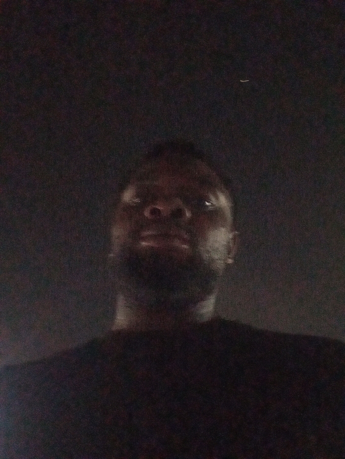 A low-angle, dimly lit photograph captures a person with dark skin. The person's face is partially illuminated, revealing facial features such as the nose, eyes, and a beard. The upper body is also visible, with dark clothing blending into the dark background. A small white line appears in the upper part of the background.