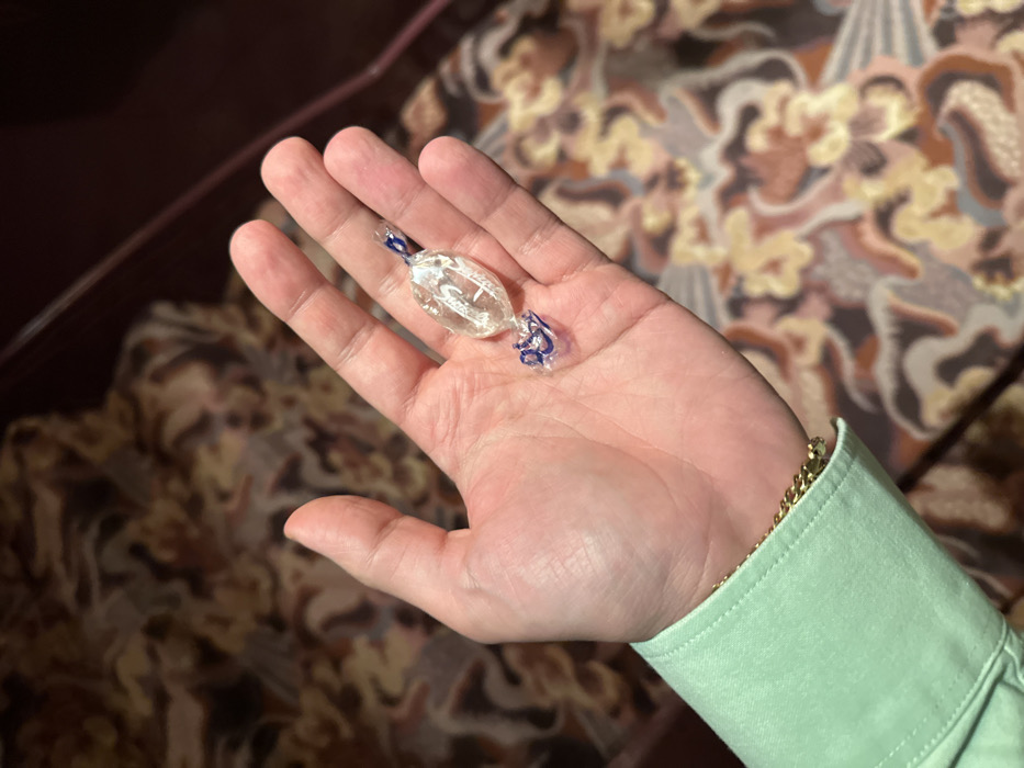 A light-skinned human hand, appearing to be an adult's, is depicted holding a single wrapped piece of hard candy in a palm-up orientation. The subject wears a light green long-sleeved shirt; a gold-colored chain or bracelet is visible on the wrist, partially obscured by the shirt cuff. The candy is oval-shaped, translucent, and appears light-colored, enclosed within a clear plastic wrapper. The wrapper is twisted at both ends, with visible blue accents, more pronounced on the lower twisted portion. Indistinct white patterns or markings are discernible within the candy itself. The background is out of focus, revealing a patterned surface consistent with upholstery or carpet, featuring abstract organic shapes in shades of brown, beige, olive green, and light blue. A dark, solid-colored surface, possibly furniture or architectural trim, is visible along the right edge of the frame. The precise location within the city of London, United Kingdom, cannot be determined from the visual evidence.