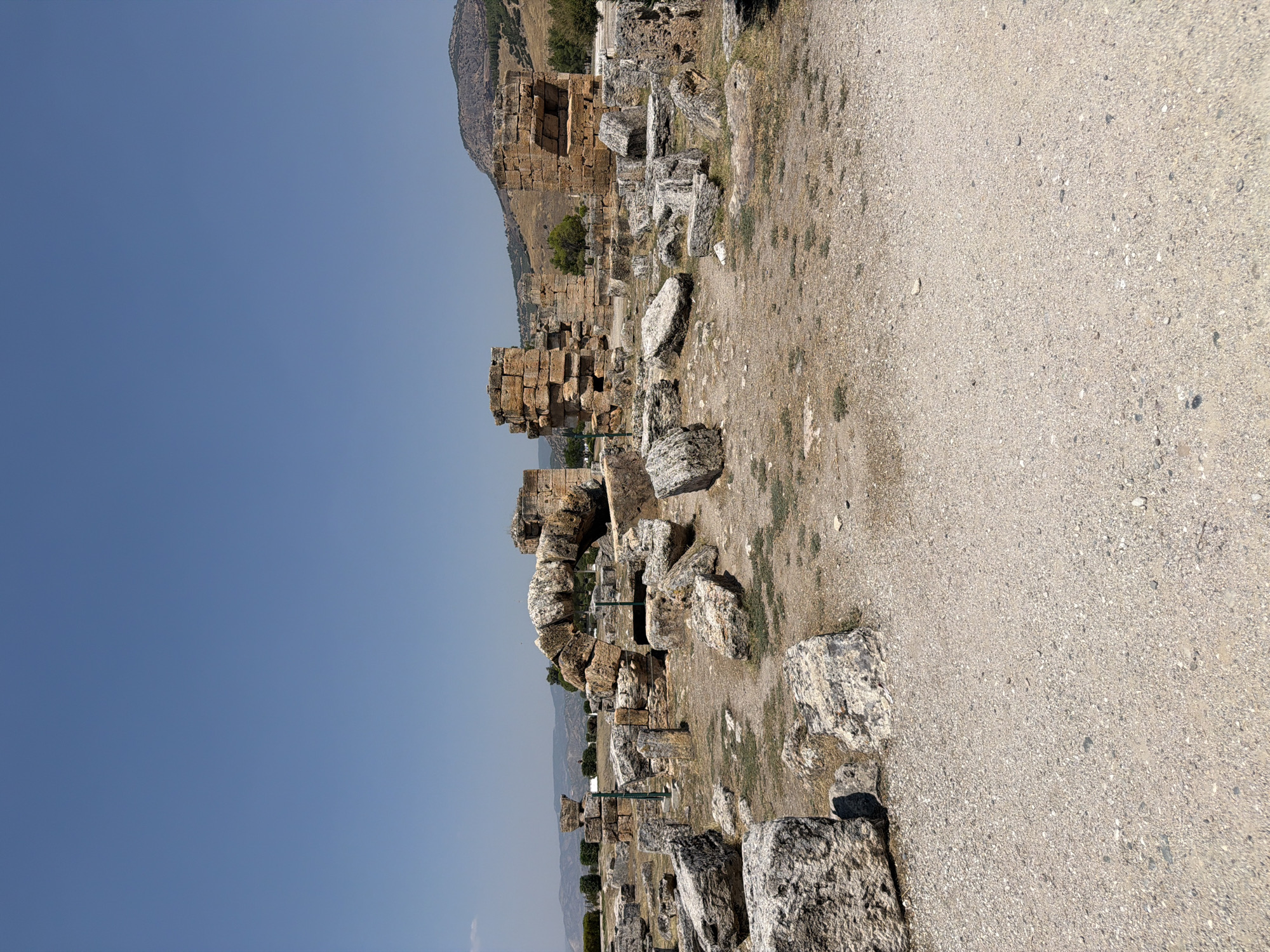 The image depicts the ruins of an ancient structure in Pamukkale, Turkey. The scene is characterized by scattered large stone blocks and fallen masonry, suggesting a historical site. A notable feature is a partially intact arched structure made of large, irregular stones, supported by upright stone pillars. In the background, more ruins, including rectangular stone structures resembling walls or columns, are visible. The ground is covered with gravel and patches of dry grass. The sky is clear blue, and distant hills with sparse vegetation can be seen. There are no people present in the image.