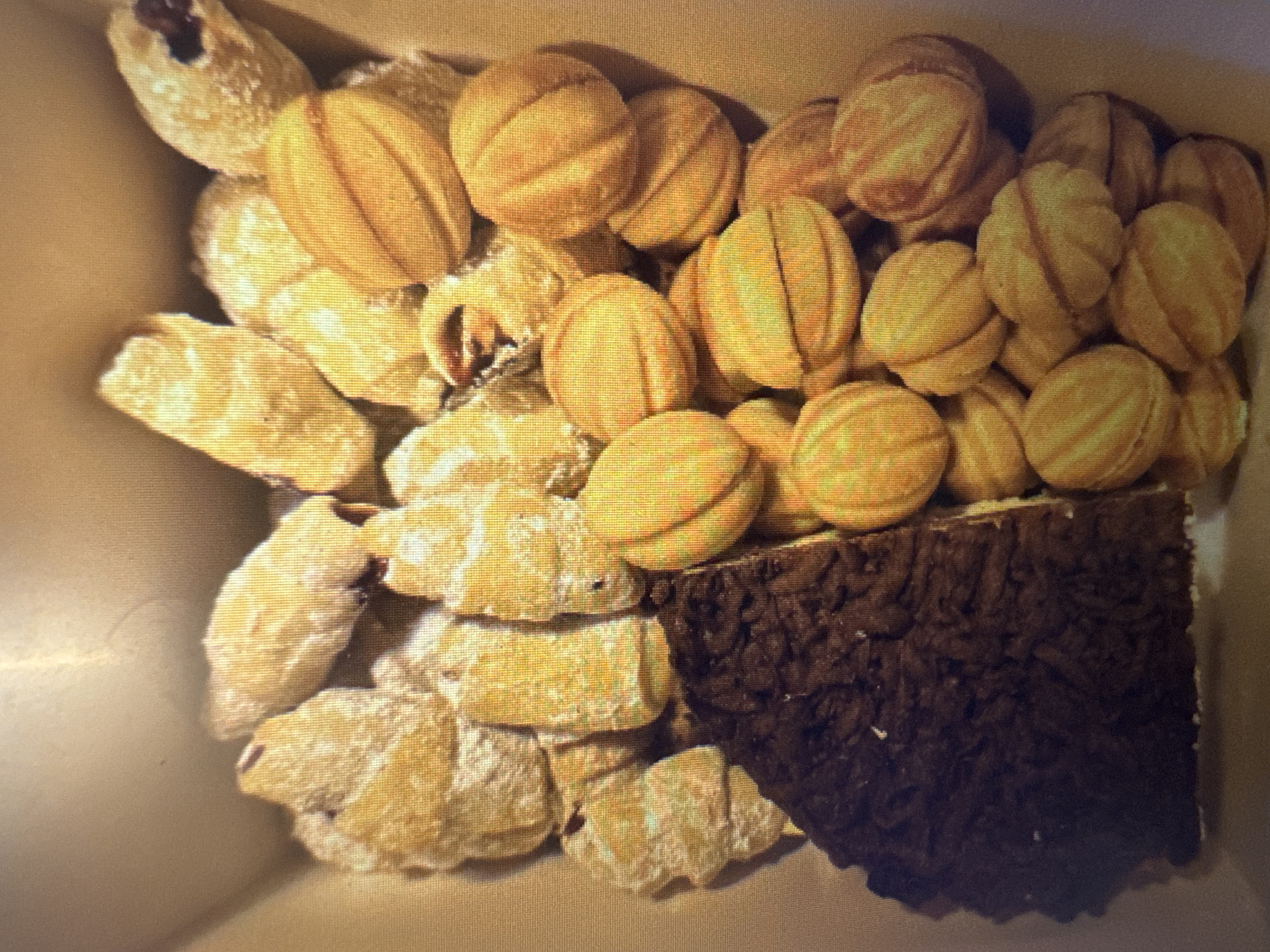 The image displays a close-up, top-down view of a collection of pastries in a light-colored cardboard box. The pastries are varied in appearance. On the left side, a large, dark brown, rectangular piece of cake or brownie is visible, with a textured, slightly crumbly surface. To the right of this, and filling the rest of the box, are numerous small, light brown, oval-shaped cookies or pastries. These cookies have a distinctive ribbed pattern on their surface, resembling halved walnuts or small buns. Some of these cookies are dusted with a white powder, possibly powdered sugar. Several of the cookies appear to be filled with a darker substance, possibly jam or cream. The overall impression is of a confectionery assortment. No specific location in Murcia, Spain is identifiable from the image itself.