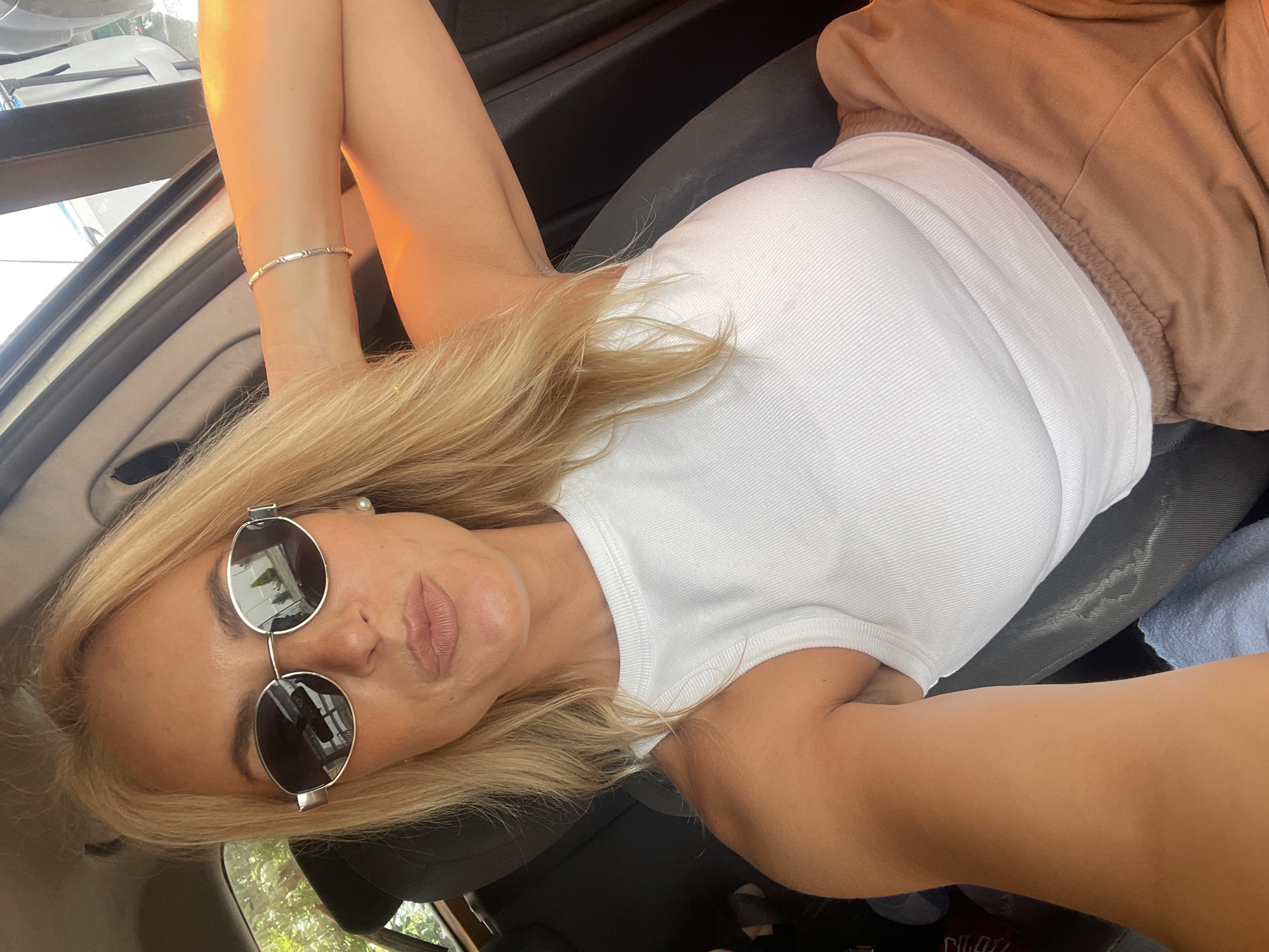 A blonde woman with fair skin and light makeup is in the driver's seat of a car, wearing sunglasses, a white sleeveless top, and light brown shorts. She is looking at the camera with her lips pursed and one arm resting on the back of her head. A bracelet is visible on her wrist. The car's interior is dark, with sunlight streaming in from the side window. Outside the car, trees and a road are visible. The location is the city of Baranovka, Russia.