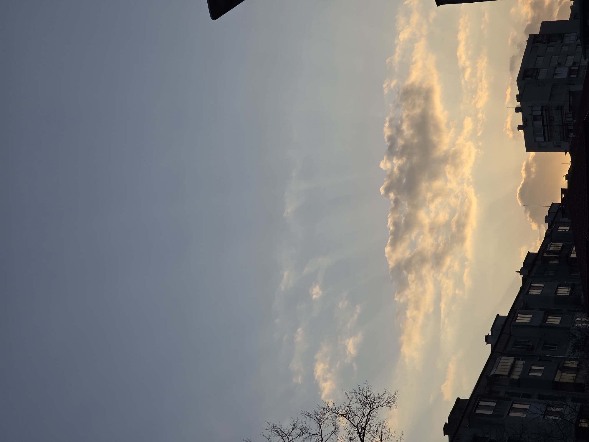 The scene depicts an urban sky over Varna, Bulgaria, at either sunrise or sunset. The upper two-thirds of the image display a sky transitioning from a light, clear blue at the zenith to warmer hues of pale orange and yellow closer to the horizon. Scattered stratocumulus clouds are present across the middle and lower sky, some illuminated directly by the low-angle sun, with distinct crepuscular rays visible radiating upwards from the horizon through the atmospheric haze. The lower third of the frame is dominated by the dark silhouettes of several multi-story residential or apartment buildings, characteristic of Eastern European urban architecture. These structures feature multiple windows, balconies, and flat roofs. On the far left, a bare, deciduous tree branch is visible in silhouette against the sky. A dark, angular object, likely a portion of a streetlamp or utility pole, is present in the upper right corner, also silhouetted. No individuals or discernible actions are visible within the frame.