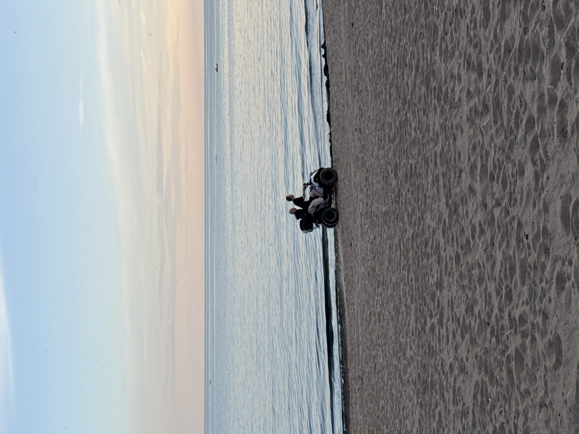 Two individuals are riding a white all-terrain vehicle (ATV) on a sandy beach. The ATV is positioned close to the water's edge. The sea is calm with gentle waves, and the sky above is a soft gradient of blue and pink, suggesting either sunrise or sunset. The beach itself is composed of textured sand. In the distance, on the water, there are a few small boats or buoys. The overall scene depicts a serene coastal setting in Zelenaya Roshcha, Russia.