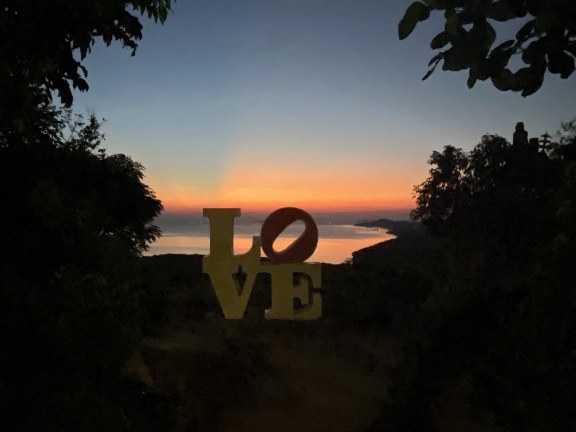 The image captures a twilight scene in Karimunjawa, Indonesia. In the foreground, silhouetted against the fading light, are the letters "LOVE" in large, yellow, three-dimensional sculptures. The letter "O" is a distinct reddish-orange ring. These sculptures are positioned on a dark, wooded hillside.
Behind the "LOVE" sculptures, the sky transitions from a deep blue at the top to hues of orange and pink near the horizon, indicating sunset or sunrise. Rays of light emanate from the horizon, illuminating a body of water, likely the sea, with islands or landmasses visible in the distance. The scene is framed by dark foliage, suggesting the image was taken from a vantage point surrounded by trees. The overall mood is tranquil and romantic, amplified by the dramatic sky and the prominent word "LOVE."