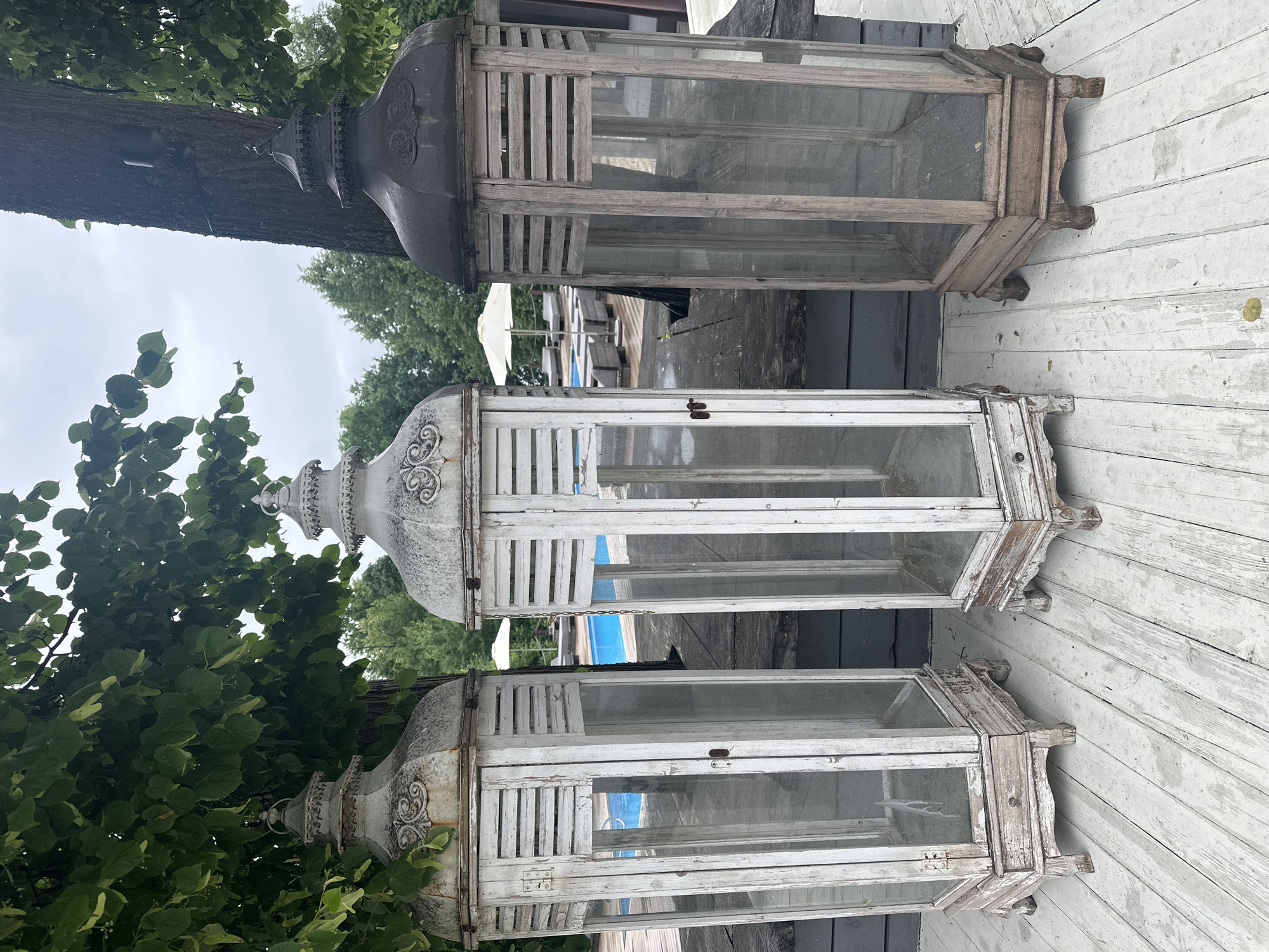 Three tall, decorative lanterns stand on a weathered wooden deck. The lantern on the left and the middle lantern are painted white with visible signs of wear and distressing, including peeling paint and some rust on the ornate tops. Both feature slatted upper sections and glass panels on their lower, hexagonal bodies. The lantern on the right has a dark metal top and a weathered wooden body with glass panels, also appearing aged. In the background, a swimming pool with bright blue water is visible, along with lounge chairs, umbrellas, and trees. The sky is overcast. The scene suggests an outdoor, possibly resort or residential, setting in a location like посёлок гидроузла имени Куйбышева, Russia. There are no people or animals in the image.