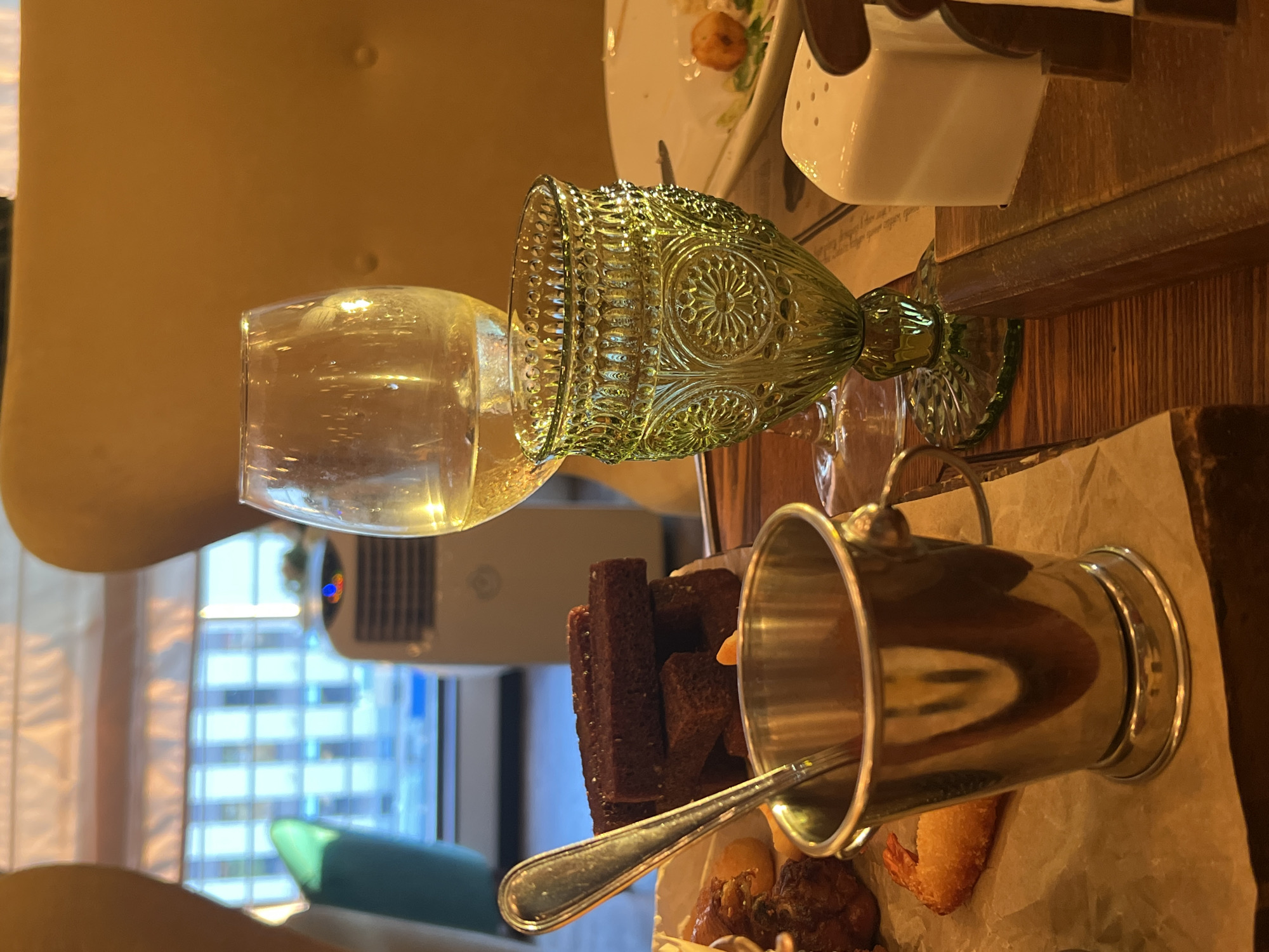 An indoor scene, likely a restaurant in Novosibirsk, Russia, features a wooden table setting. In the foreground, a silver bucket-shaped container with a spoon inside holds a red sauce. To the left, a pile of dark rectangular croutons is visible. Behind the croutons, a fried shrimp is partially visible on a piece of parchment paper. In the center right, a decorative green glass goblet stands next to a wine glass filled with a pale yellow liquid. A white salt shaker is situated to the right of the goblets. In the background, a large window reveals a building with illuminated windows, and a white portable air conditioner unit is visible. The overall lighting is warm, suggesting an evening or interior lighting.