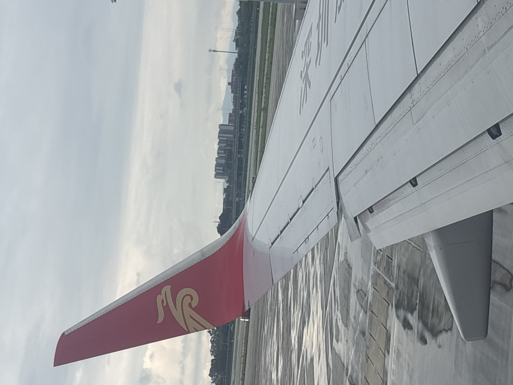 The image, taken from an airplane window, showcases the wing and tail fin of an aircraft against a cloudy sky. The tail fin is predominantly red with a golden emblem resembling a bird or phoenix. The wing is white with visible panel lines and some markings. In the background, an airport tarmac with puddles of water is visible, reflecting the overcast sky. Further beyond the tarmac, a city skyline with buildings and some trees can be observed. The scene suggests the aircraft is either on the ground at an airport or in the process of taking off or landing. The presence of buildings and infrastructure in the background indicates an urban or semi-urban environment. Based on the airline's livery, the aircraft likely belongs to China Southern Airlines, which uses a red tail fin with a golden flower emblem. The city in the background is identified as Baiyun District, China, which is known for housing Guangzhou Baiyun International Airport, a major hub for China Southern Airlines.