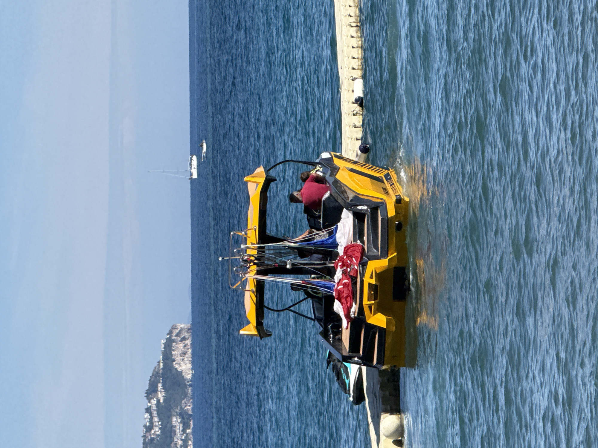 In Budva, Montenegro, a bright yellow speedboat with a canopy is docked next to a floating platform. The boat has people on board and equipment for parasailing, including colorful parachutes. A jet ski is also attached to the back of the boat. The water is a deep blue, and in the background, a coastal town is visible on a hillside, with sailboats in the distance. The scene suggests a recreational activity, possibly parasailing tours, taking place on a clear, sunny day.