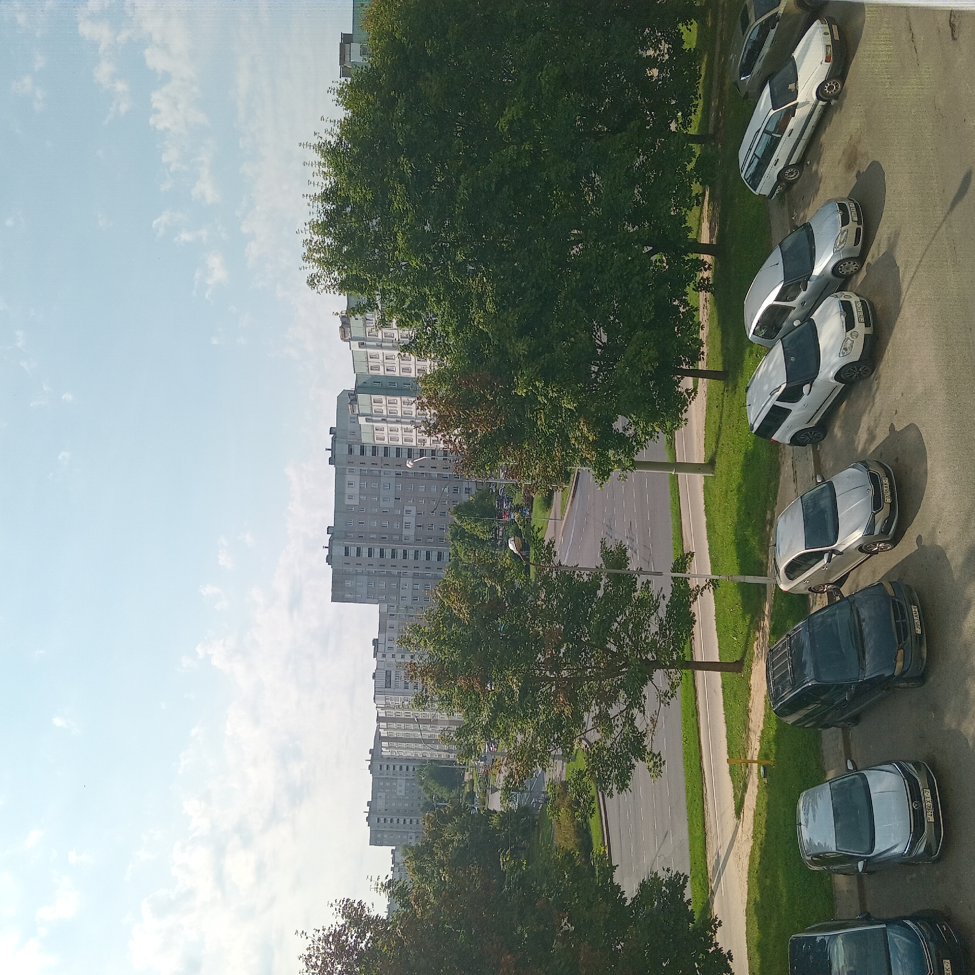 The image depicts a view of apartment buildings and a street in Minsk, Belarus. In the foreground, there is a parking lot with several cars. From left to right, the visible cars include a dark blue sedan, a silver Volkswagen sedan, a dark minivan, a silver Skoda sedan, a white Skoda Yeti SUV, a white Volkswagen Passat wagon, and a dark SUV. The cars are parked on a paved surface, with grass and trees bordering the parking lot and street.
The street is a two-lane road with white dashed lines marking the lanes. Across the street, there is a grassy area with trees and more apartment buildings in the background. The apartment buildings are multi-story, typical of Soviet-era architecture, and appear to be residential buildings. The sky is partly cloudy, with a clear blue sky dominating the upper portion of the image. The overall scene is a residential area of Minsk, likely a microdistrict characterized by high-rise apartment blocks and surrounding green spaces.