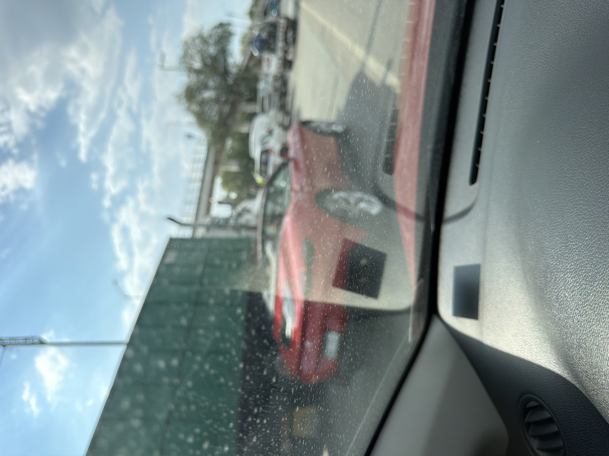 The image, taken from inside a vehicle, shows a red car visible through the passenger-side window, with other vehicles indistinctly visible in the background. The sky is partly cloudy. The dashboard of the vehicle the photo was taken from is in the foreground. There's nothing in the image to confirm the location as San Bernardino Tlaxcalancingo, Mexico.