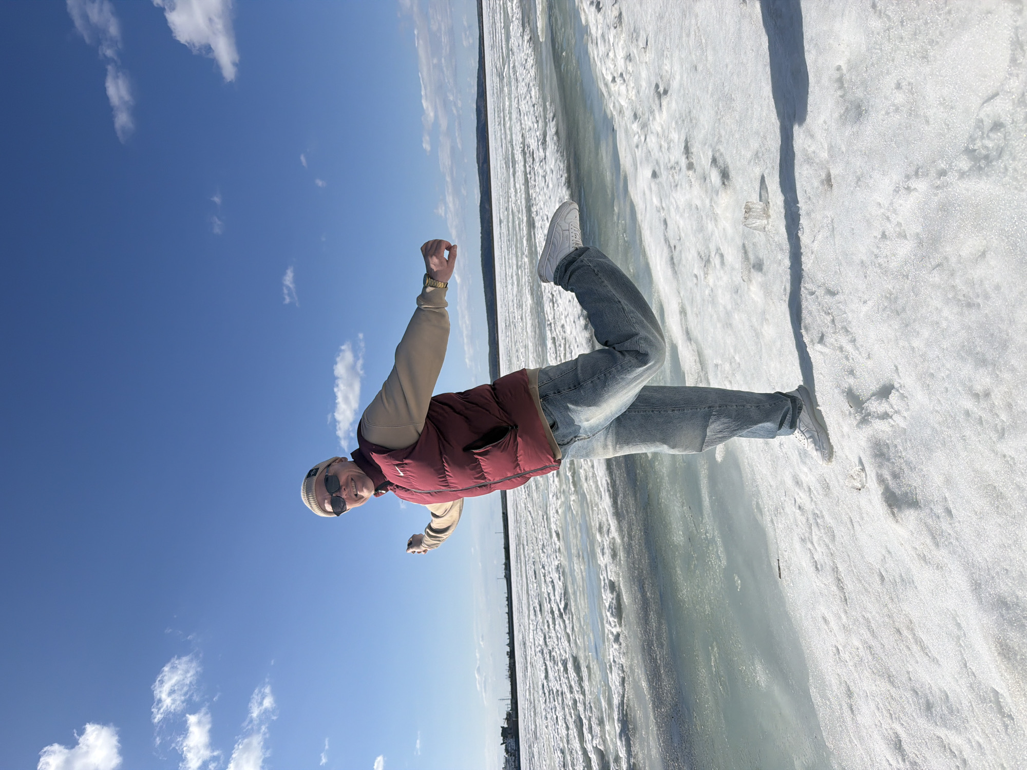 A man stands on a snow-covered surface next to a frozen body of water on a bright, sunny day. The man is wearing a light-colored beanie, sunglasses, a tan long-sleeved shirt, a red vest, light blue jeans, and white sneakers. He is raising his arms in a gesture of triumph or excitement. The snow appears to be on a frozen body of water. In the distance, across the water, are buildings which place this in an urban location in Chaikovsky, Russia. The sky is blue with scattered clouds. A shadow cast by the subject is visible on the snow. A clear container sits in the snow next to the shadow.