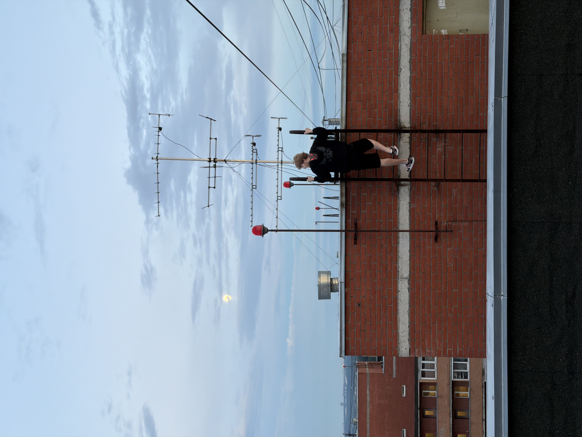 The image shows a person on a rooftop in Khimki, Russia. The person, wearing a black hoodie and shorts, is doing a pull-up on a ladder attached to the side of a brick building. The sky is cloudy, with the moon visible in the distance. The time of day appears to be late afternoon or early evening. The overall mood is one of quiet activity and urban exploration.