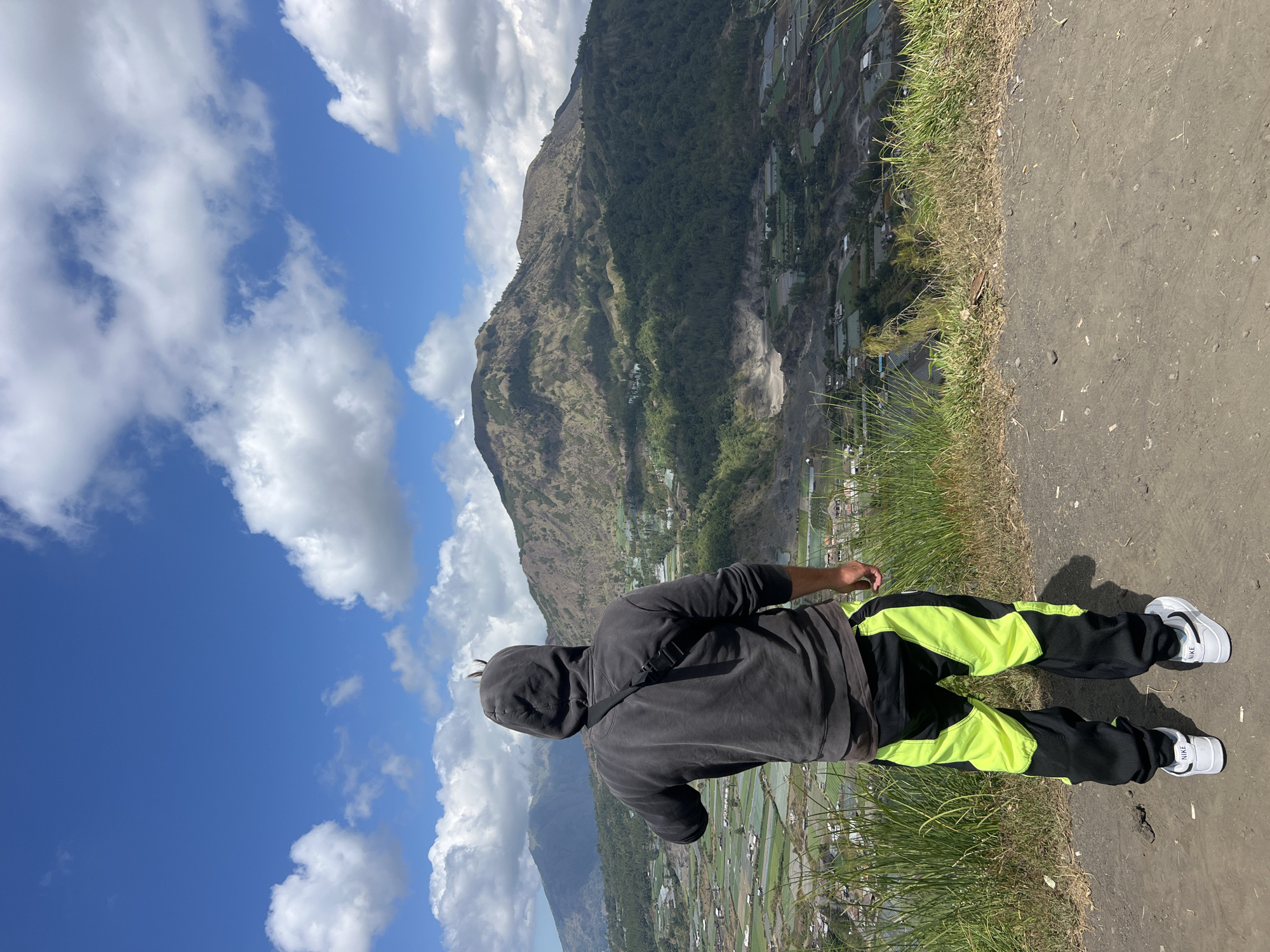 A person wearing a black hooded sweatshirt and black and neon green athletic pants stands on a dirt path, looking out over a valley in Kintamani, Indonesia. The person is facing away from the camera, with their left arm outstretched. A black cross-body bag is slung over their right shoulder. In the background, a large, verdant valley unfolds, featuring agricultural fields, scattered buildings, and the slopes of a volcanic mountain. The sky above is a vibrant blue, dotted with fluffy white clouds. The overall scene depicts a panoramic view of the Kintamani region, showcasing its natural beauty and agricultural landscape.