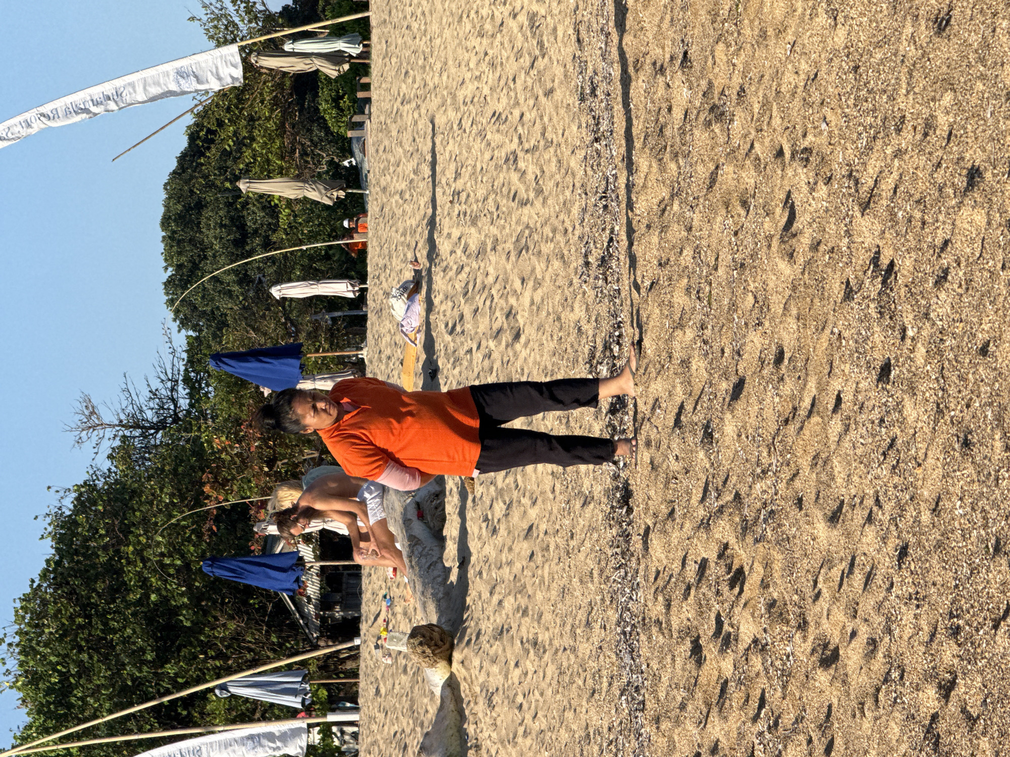 The image depicts a beach scene in Denpasar, Indonesia, with several people and beach amenities present. In the foreground, a person wearing an orange t-shirt and black pants stands with their hands behind their back, looking down. The beach is sandy, with some debris scattered across it. In the mid-ground, two people are seated on a log. One is a man with long hair, wearing white shorts, and the other is a woman. Behind them, several blue and tan beach umbrellas are visible, along with a small building structure. Further in the background, lush green trees form a backdrop against a clear blue sky. White flags on poles are also present, indicating the location is likely a public beach. The lighting suggests it is late afternoon or early morning, with long shadows cast by the objects on the sand.