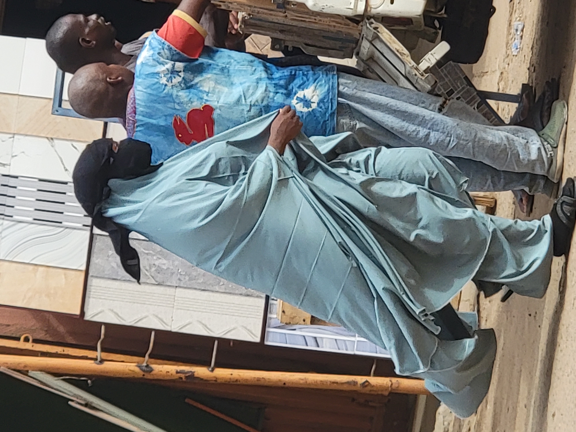 The image shows three individuals in what appears to be an outdoor setting, potentially a marketplace in Maiburiji, Nigeria. The primary subject is wearing a light blue, full-body garment with a dark head covering, positioned in a seated or squatting posture. This individual is partially obscured by the garment they are holding. Behind them, two men are visible, one wearing a blue patterned shirt with red accents and the other wearing a plain grey shirt. The setting includes building materials, potentially tiles, displayed on a rack, and a glimpse of a street or sidewalk.