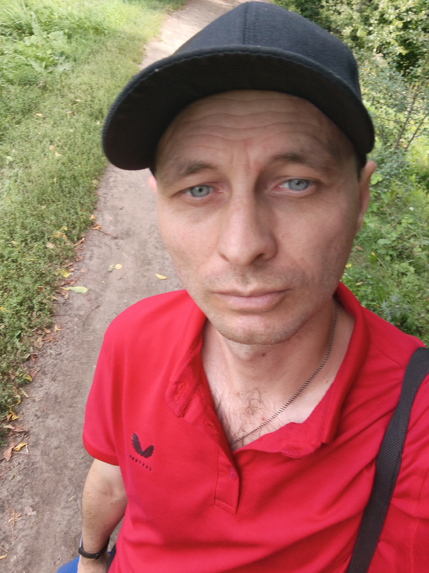 A medium shot selfie shows a man with fair skin, light blue eyes, and a short haircut. He is wearing a black baseball cap, a red polo shirt, and a silver chain necklace. The man appears to be walking outdoors, with a dirt path and green foliage visible in the background. The scene suggests a casual outdoor setting, potentially in a park or trail. There are no other people or discernible objects of significance in the frame. The lighting is natural, indicating daytime. The image quality is clear, allowing for detailed observation of the man's features and attire. The location is identified as Mokshan, Russia.