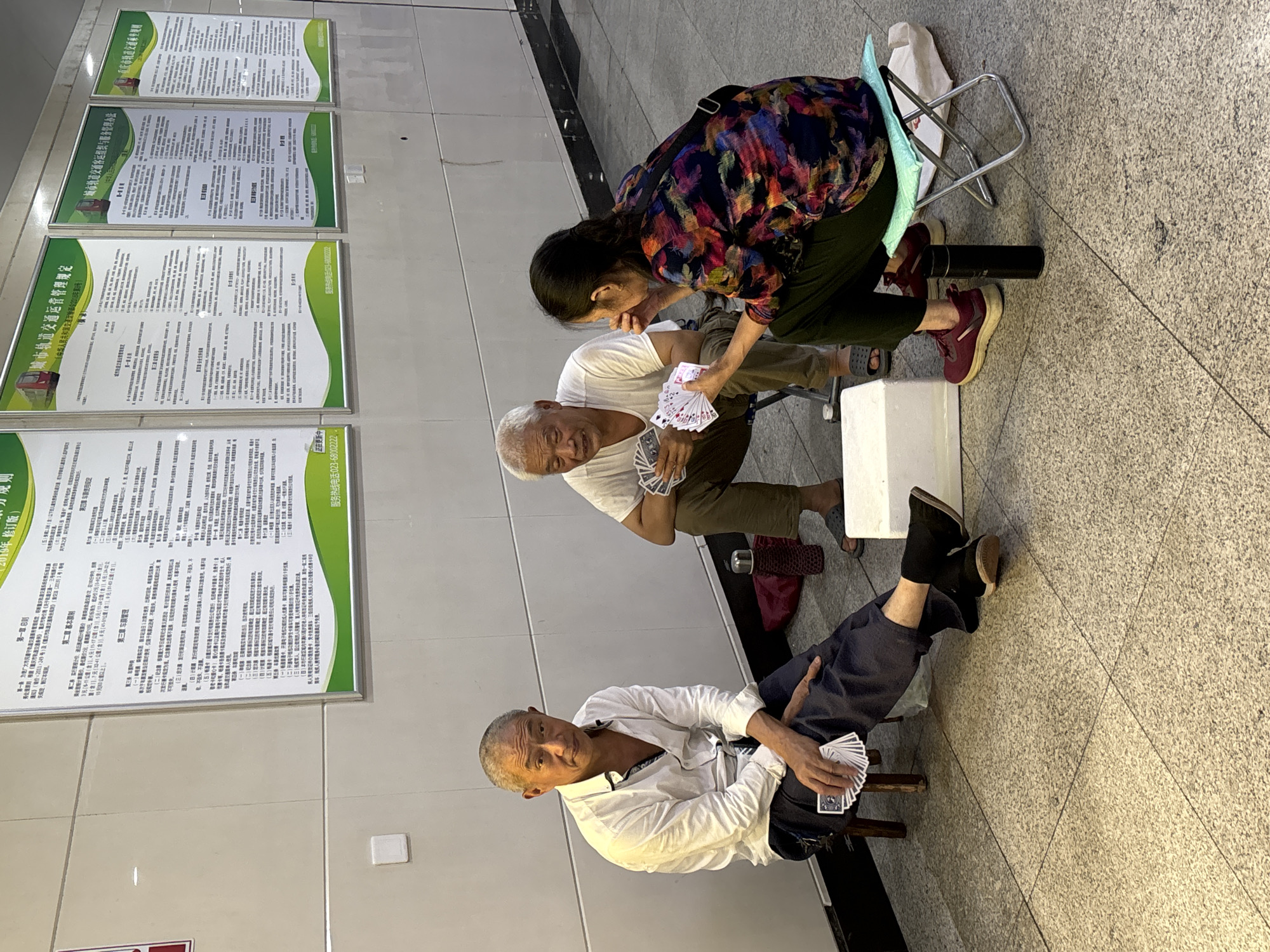 A photograph taken in an urban environment shows three elderly individuals sitting on the ground playing cards. Two men and one woman are present. The man on the left is seated on a stool and wearing a white shirt and dark pants. The man in the middle is seated on the ground in dark pants and a white t-shirt. The woman on the right is seated on a small folding stool, wearing a colorful patterned shirt and dark pants. They are all holding cards. The environment appears to be an indoor public space, possibly a waiting area or hallway, with tiled flooring and large posters or signs mounted on the wall behind them. There are various objects on the ground, including a thermos, a white cooler, and bags.