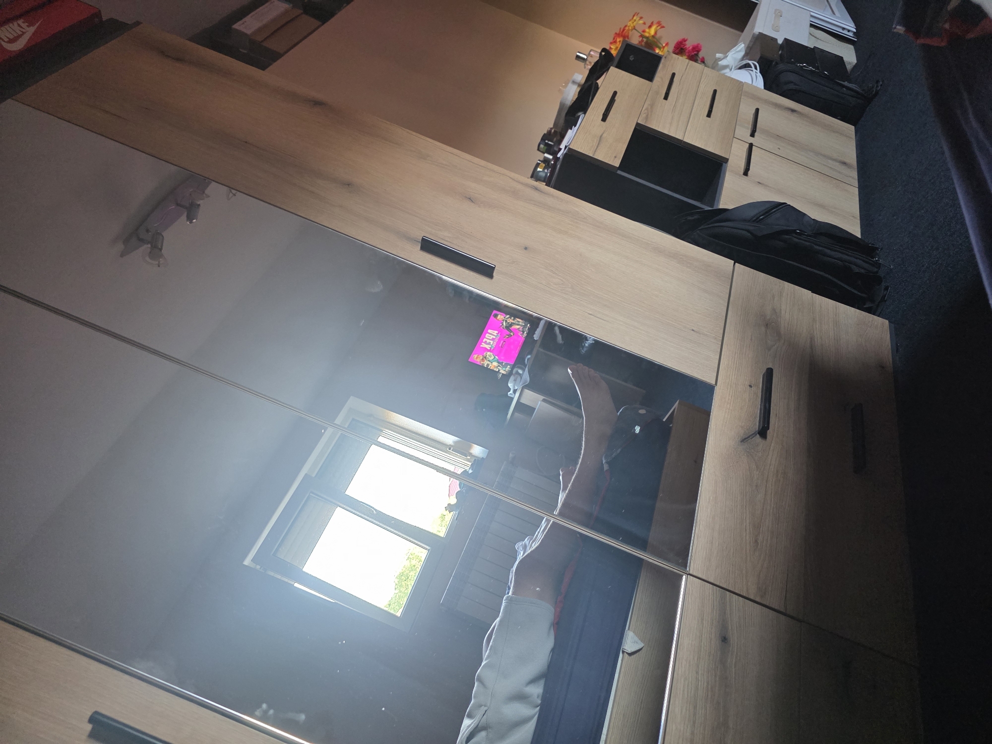 The image shows a bedroom interior, possibly in Cherven Bryag, Bulgaria, reflected in a mirrored wardrobe. The wardrobe has light wood-grain doors. The reflection shows a window with bright daylight coming through, indicating daytime. Reflected in the mirror is also a person lying on a bed with their legs raised. A book with a pink cover is visible. Next to the wardrobe is a shelving unit with black and wood panels, topped with personal items and what looks like backpacks. A red Nike shoebox is visible on the top shelf. The background is blurry and dark.